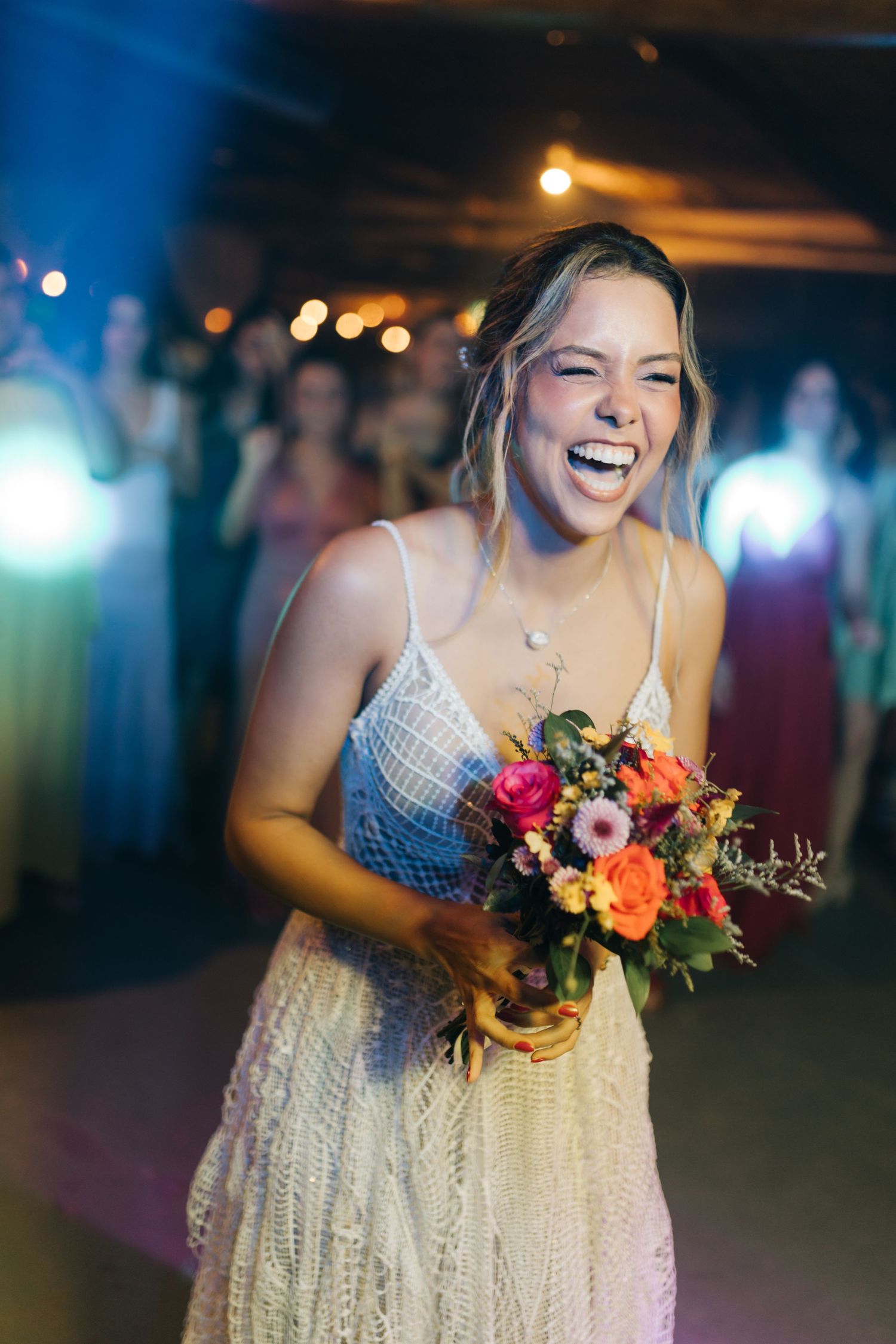 Person in white lace dress holds vibrant bouquet while laughing at celebration.