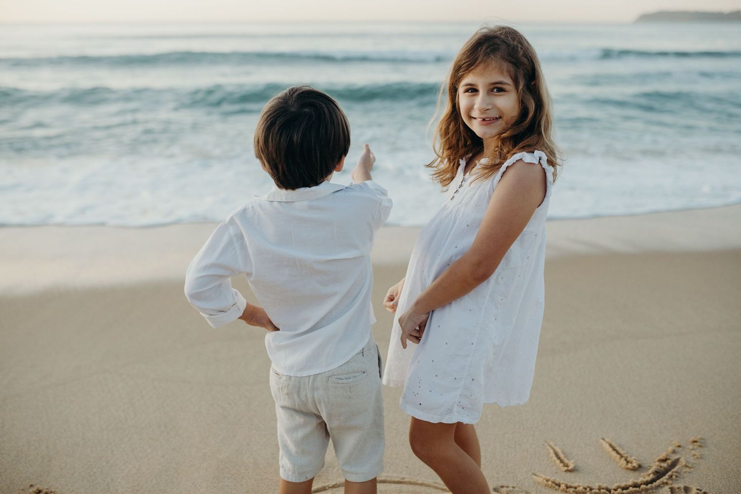 Children in white outfits enjoying a playful moment together on the beach at sunset.