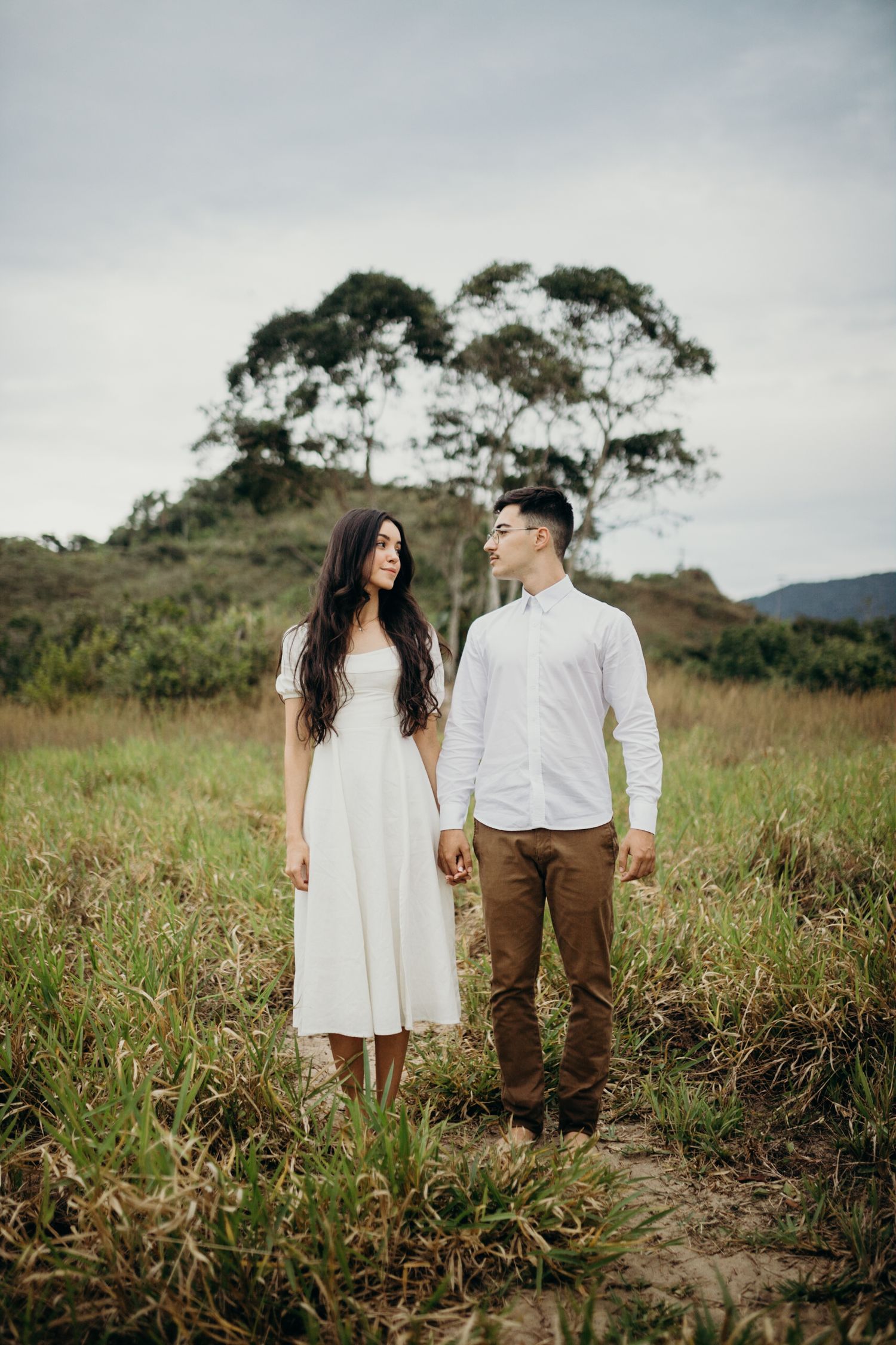 A couple in coordinated white and brown outfits pose together in a scenic hilltop meadow.