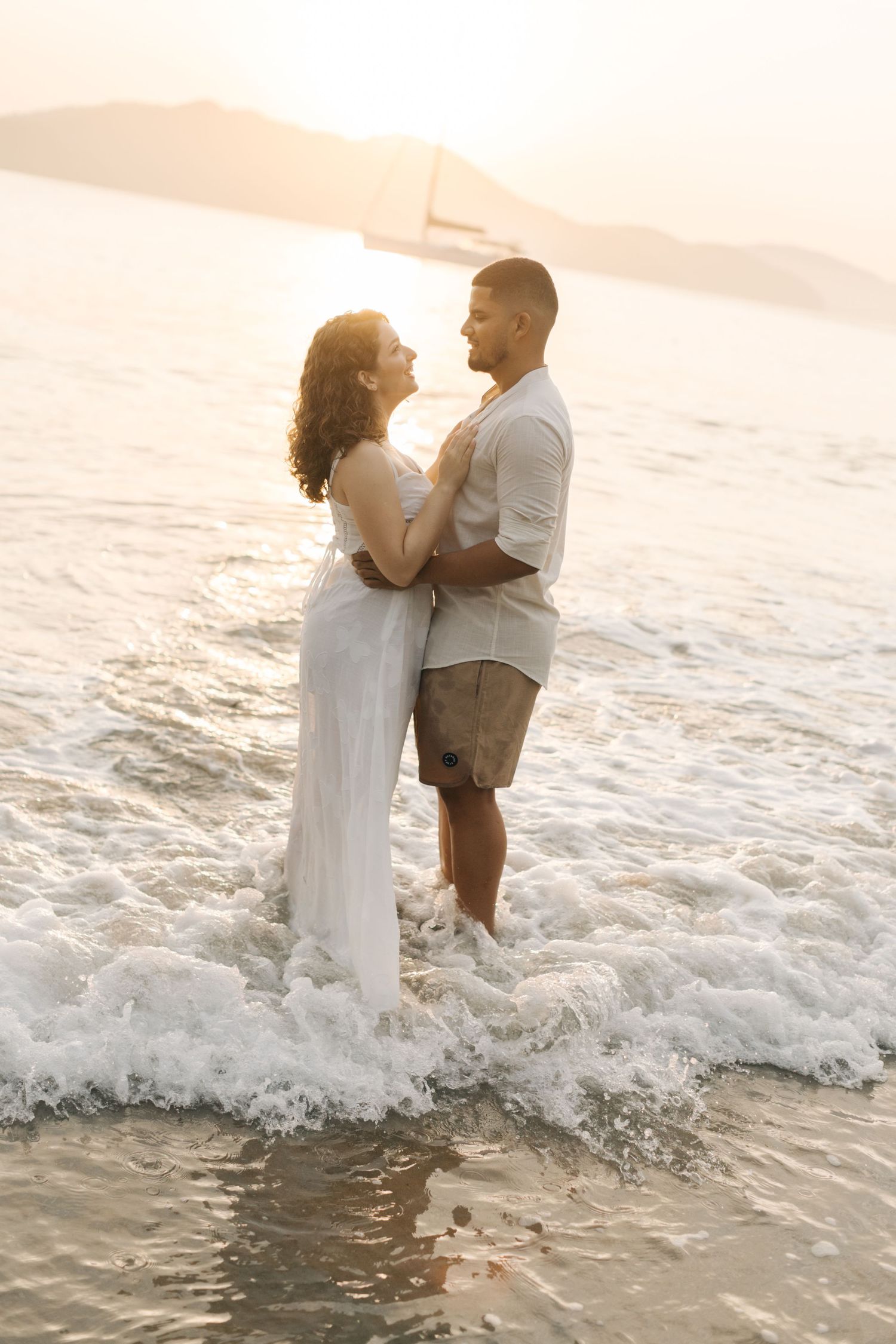 A couple in white clothing embraces while standing in ocean waves during sunset.