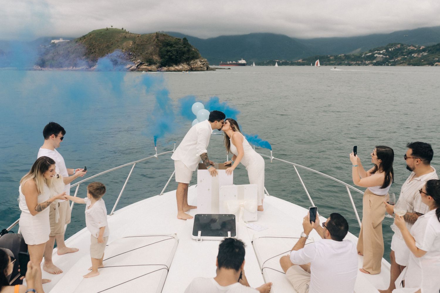 Guests create blue smoke effects during a celebration on a white yacht deck with mountains in the distance.