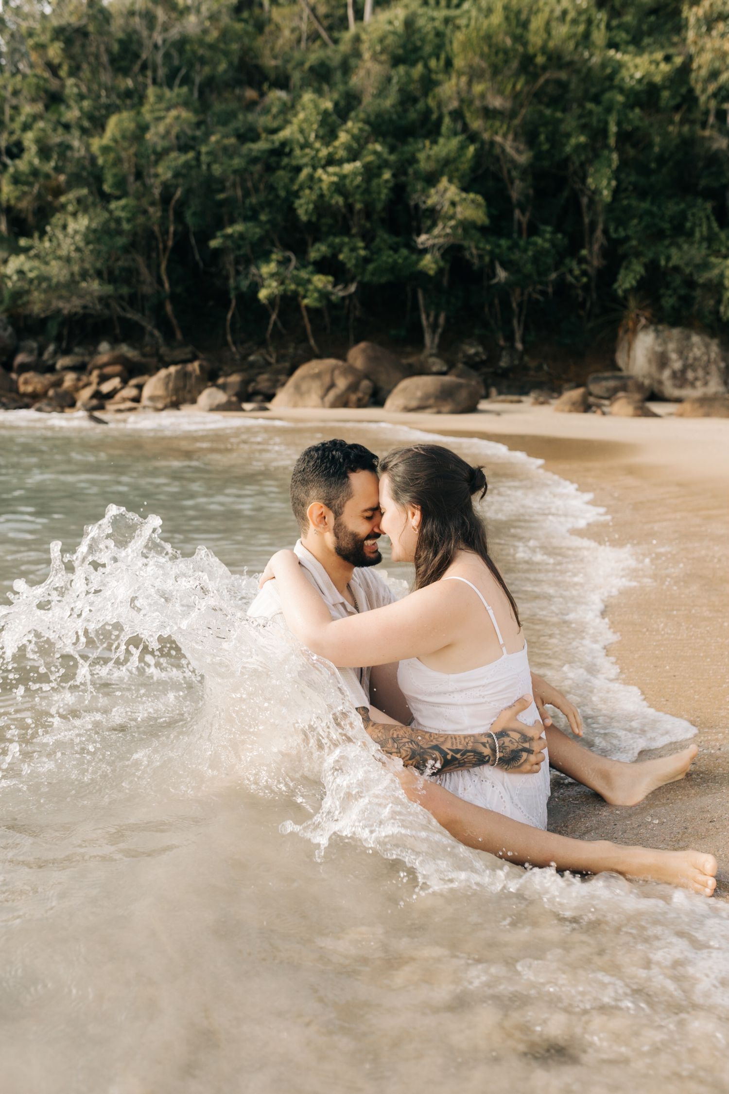 A playful couple splashes in the surf at a scenic beach with rocky outcrops.