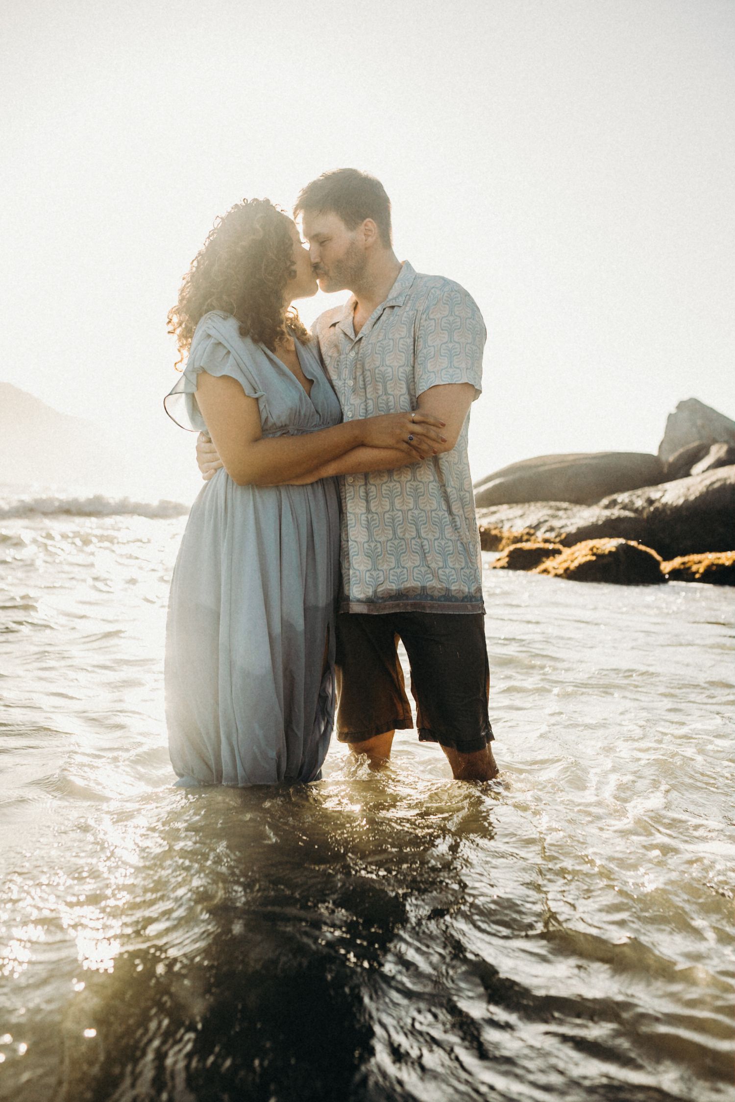 Romantic couple embracing in ocean waves at sunset on rocky beach with mountains in background.