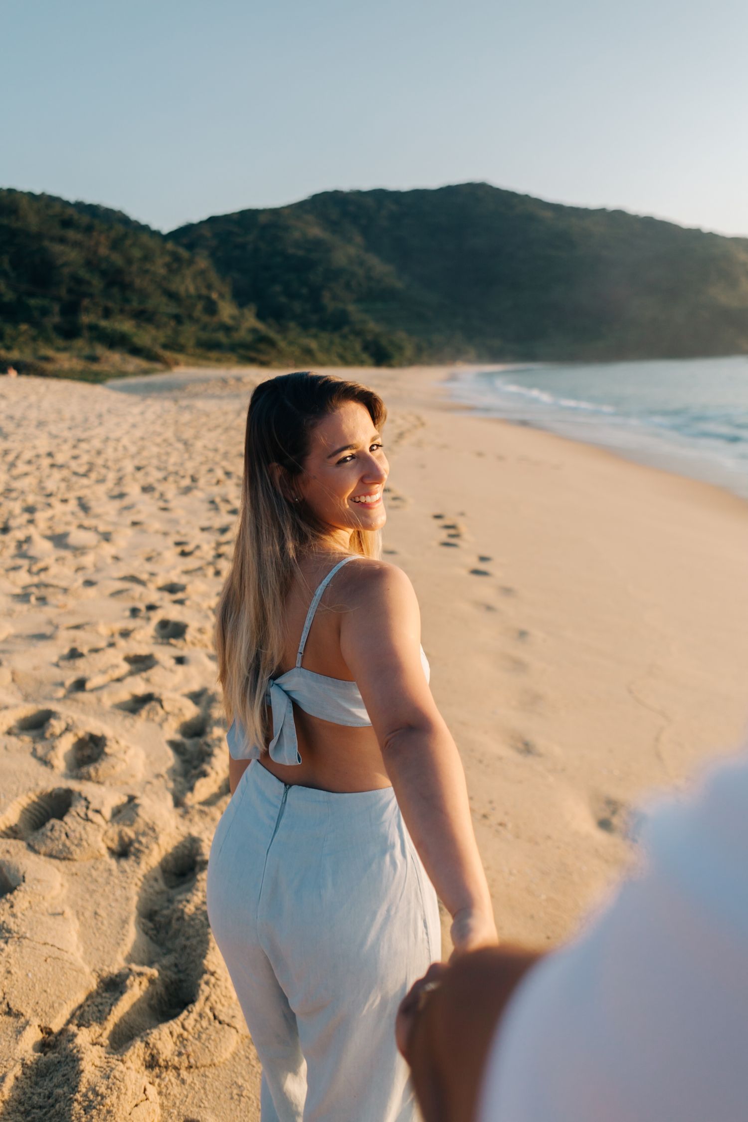 Woman in white attire standing on sandy beach looking over shoulder at sunset.