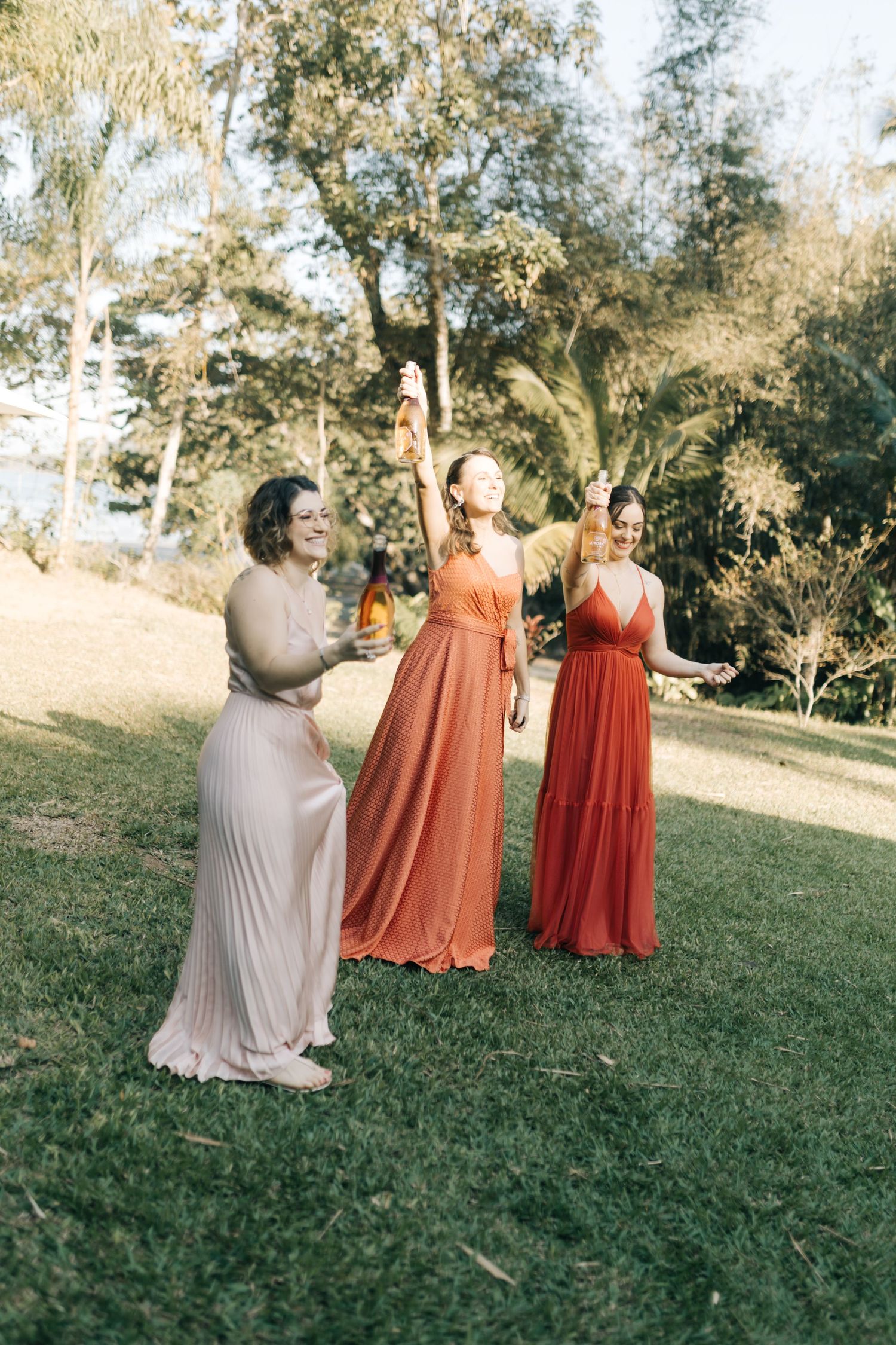 Three bridesmaids in flowing pink and coral dresses laugh together while holding drinks on a grassy lawn.