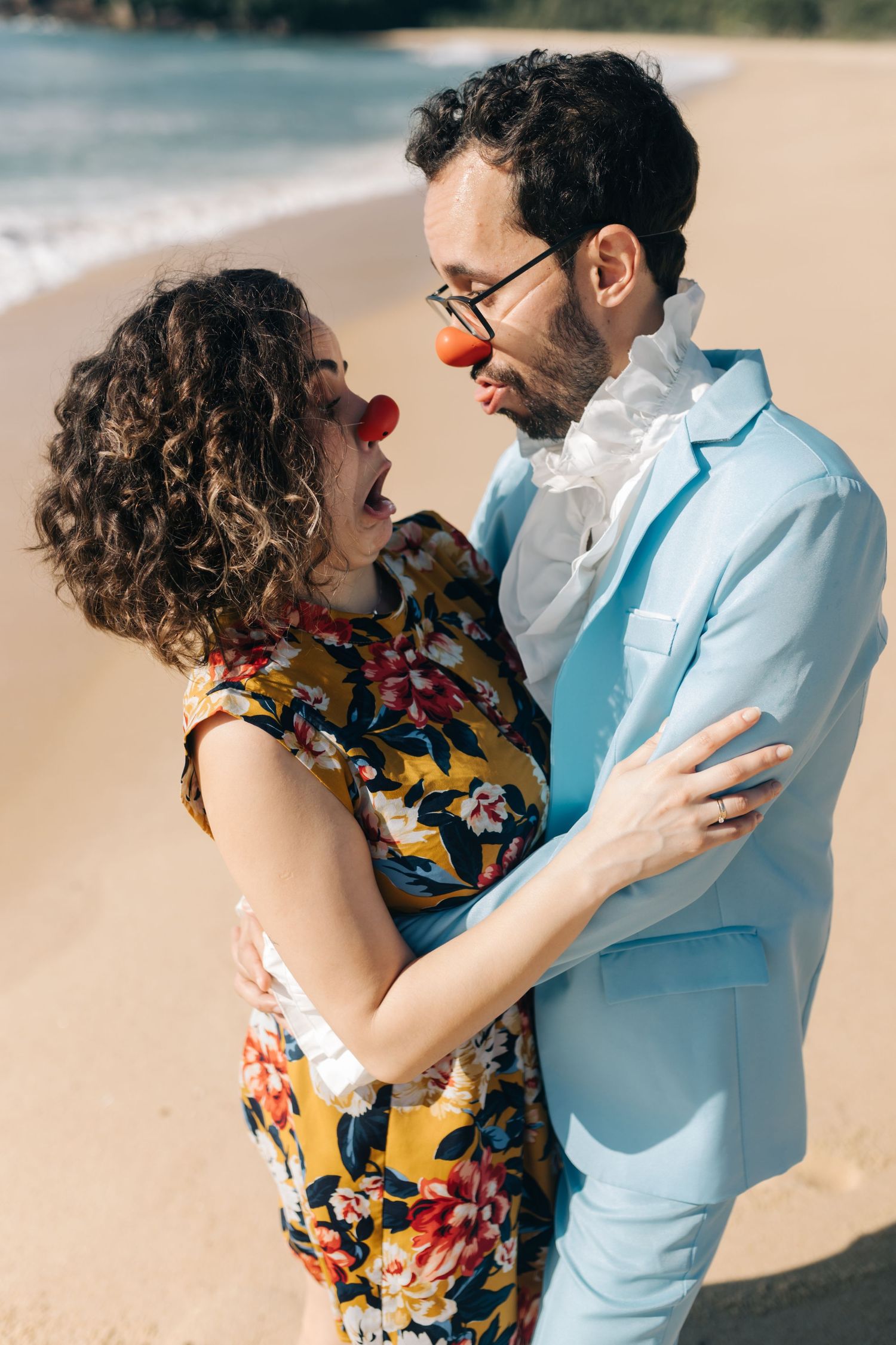 A couple in contrasting outfits share a romantic moment while wearing red clown noses on the beach.