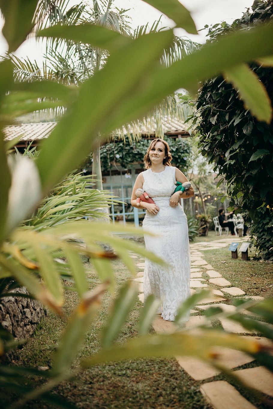 A lace-dressed figure stands on a garden path surrounded by tropical foliage and palm trees in an outdoor setting.