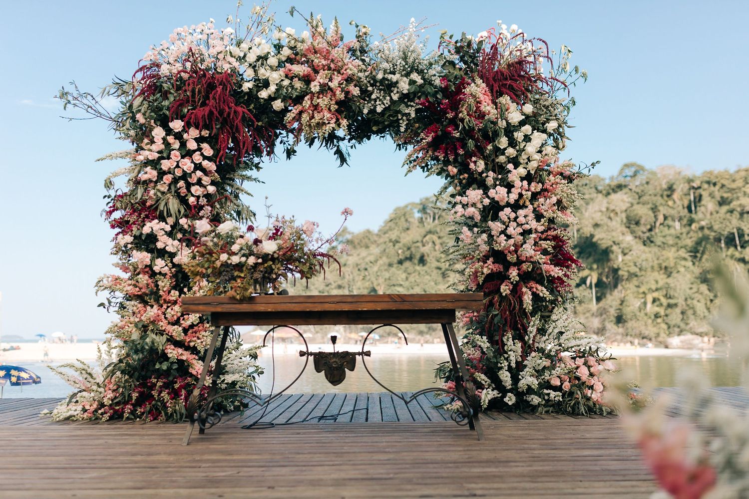 Romantic floral arch with pink and burgundy blooms against a waterfront backdrop.