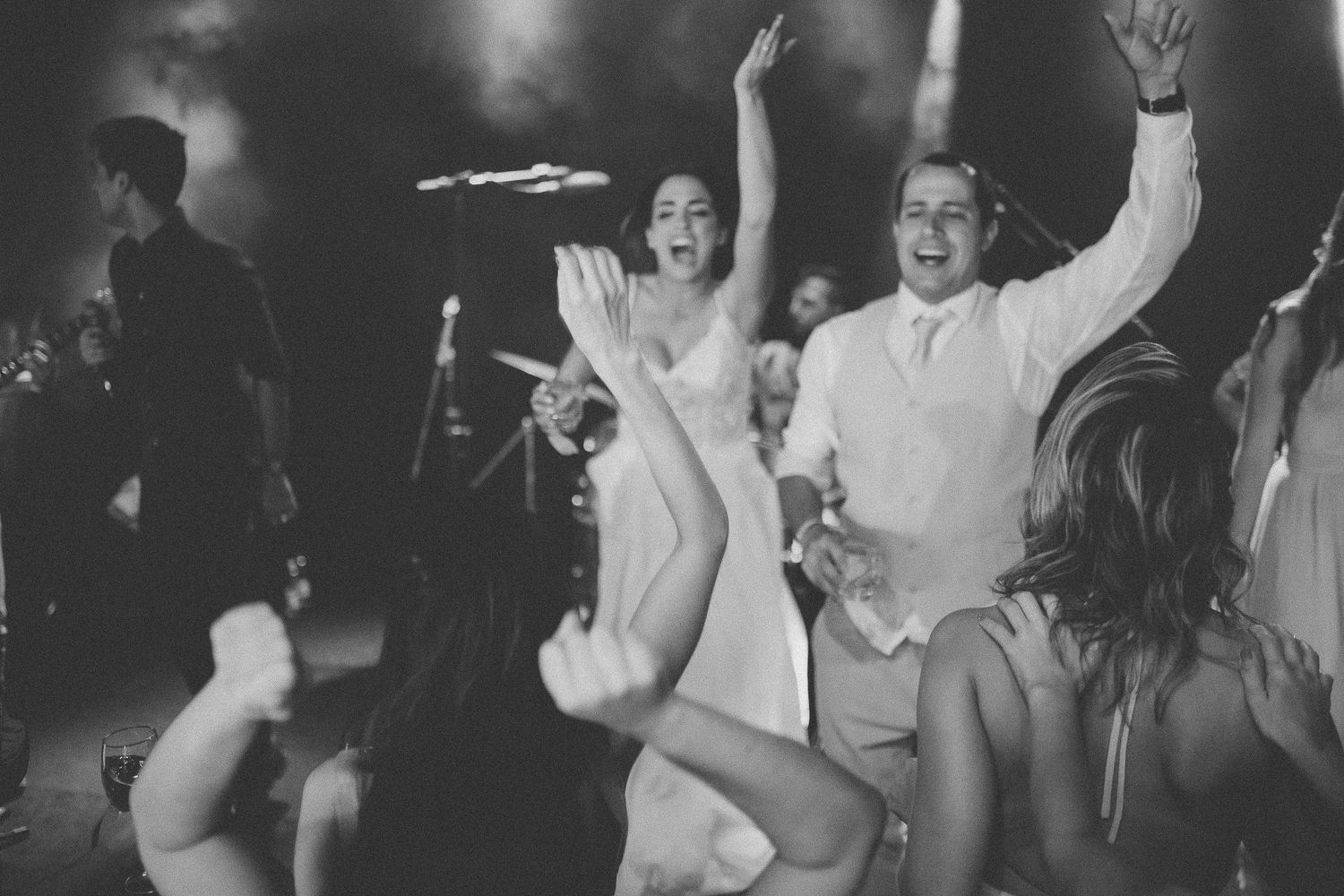 Black and white photo of enthusiastic wedding guests dancing and celebrating with arms raised.