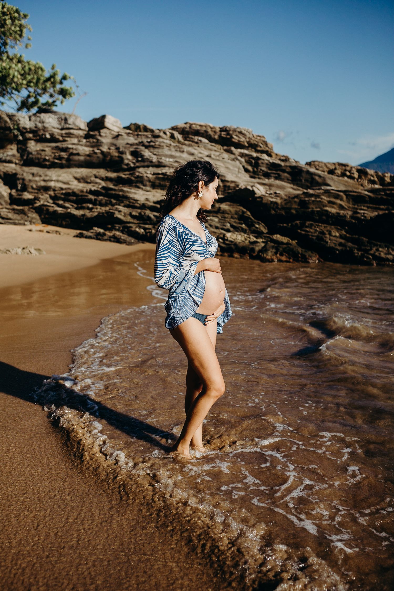 A maternity beach photo with waves washing over feet against rocky cliffs.