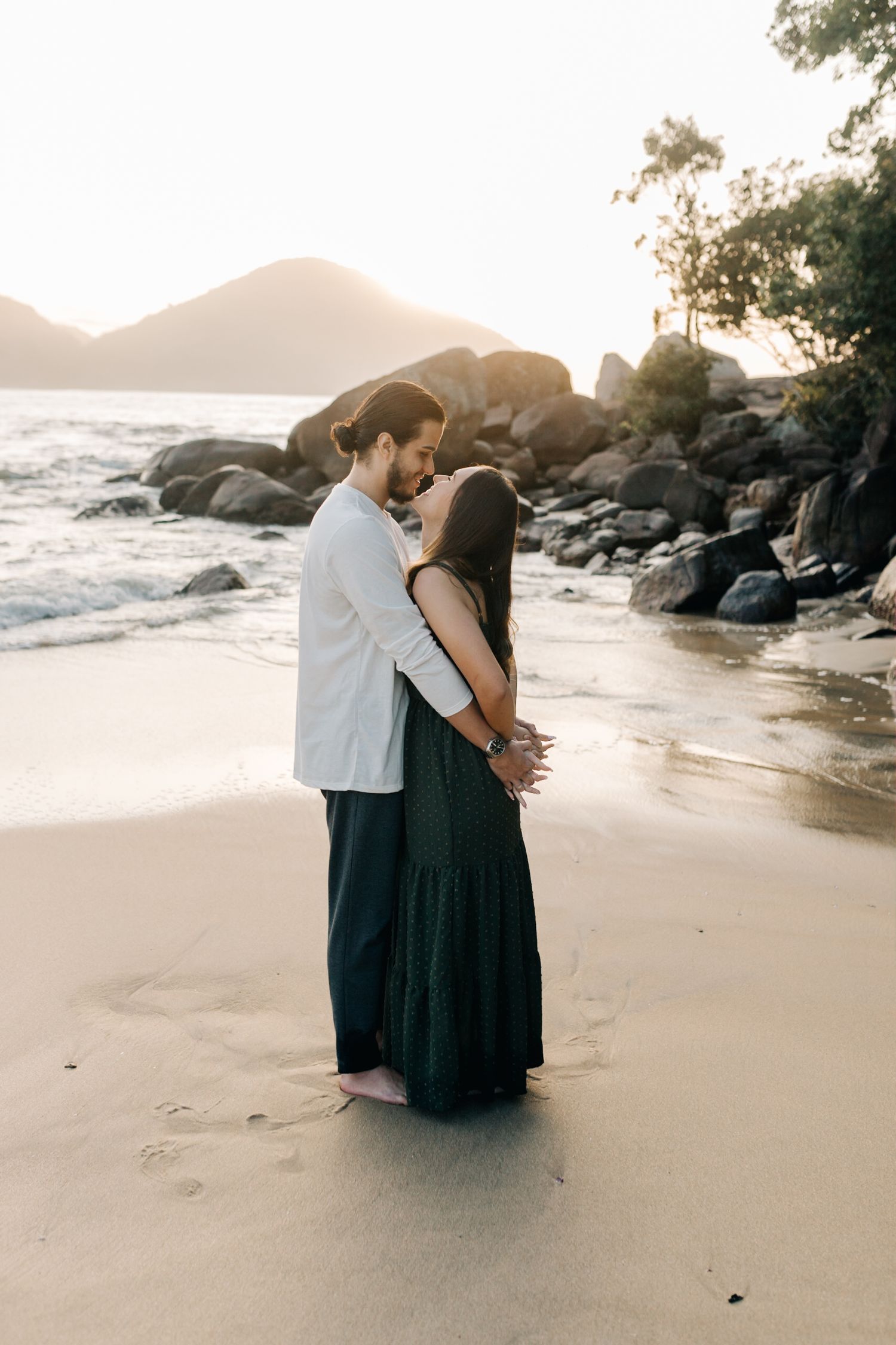 Couple shares intimate moment on beach at sunset with rocky coastline in background.
