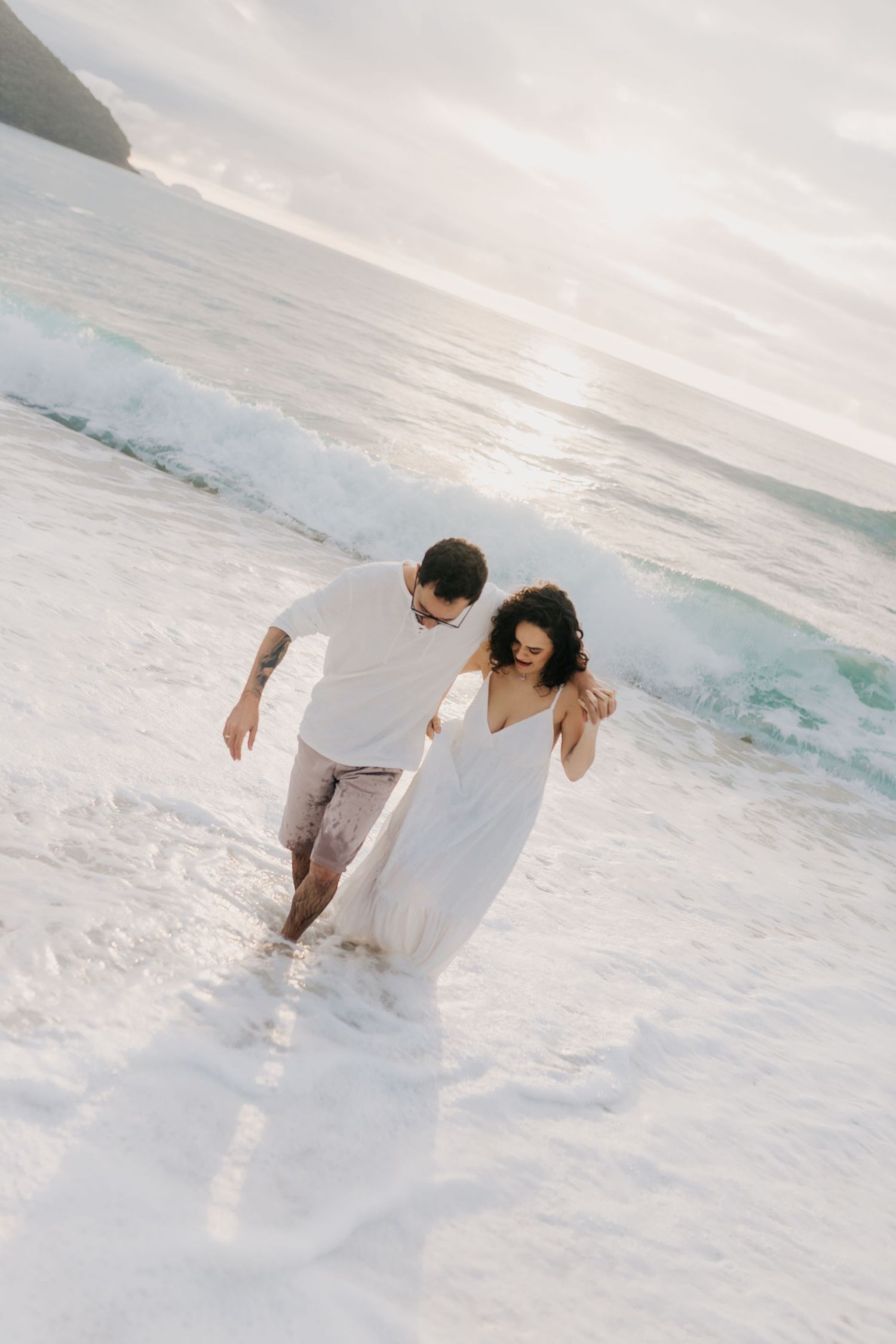 A couple walks through ocean waves wearing white and beige on a scenic beach.