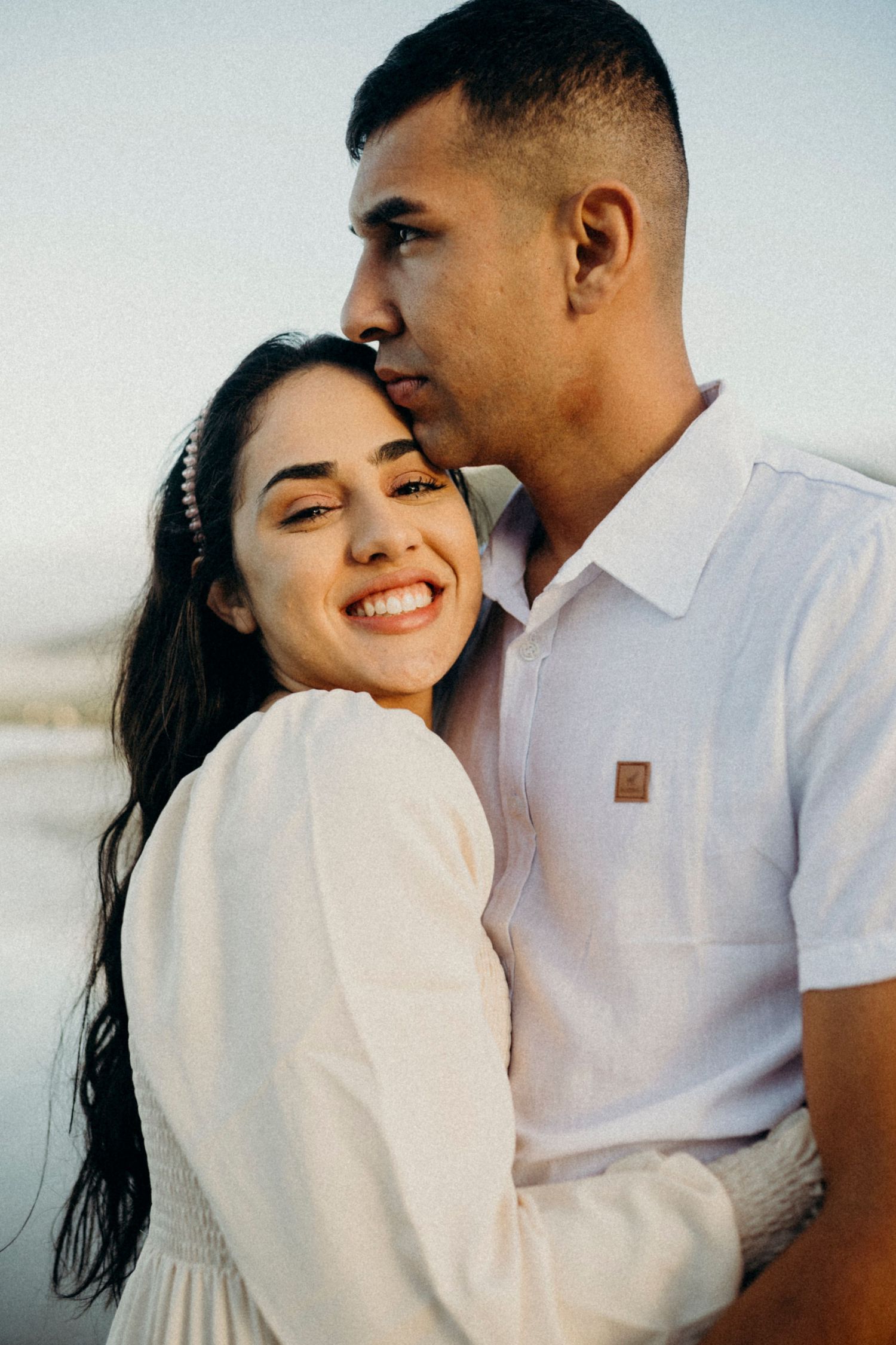 A loving couple in white clothing shares an intimate moment at the beach.