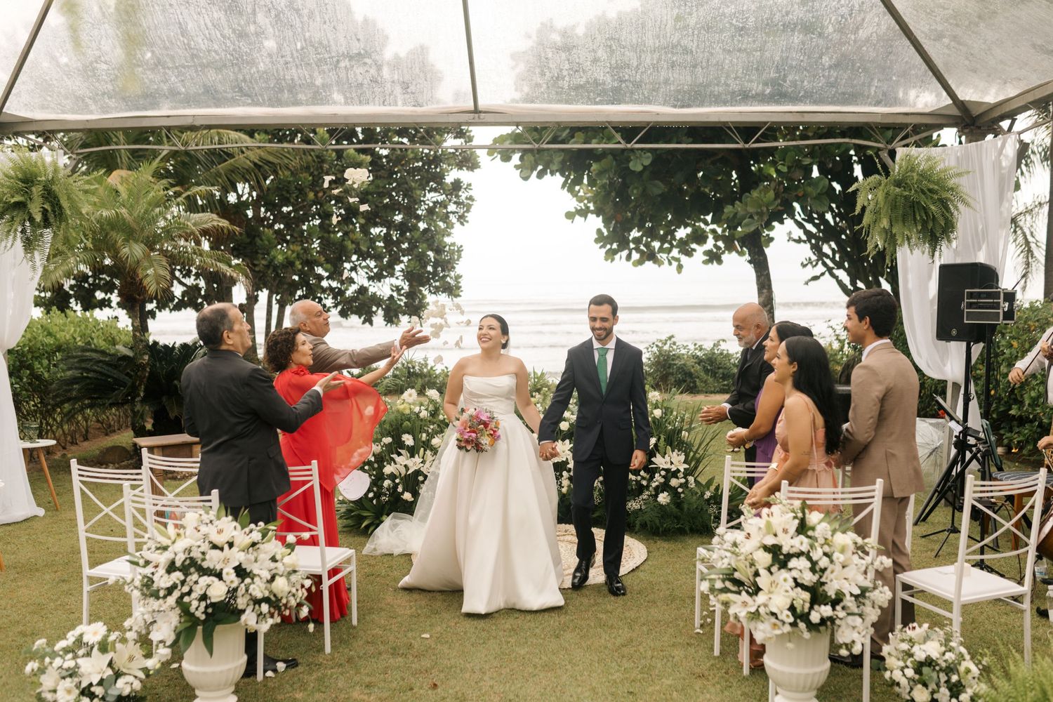 An outdoor wedding ceremony under a white tent with floral decorations and seated guests.