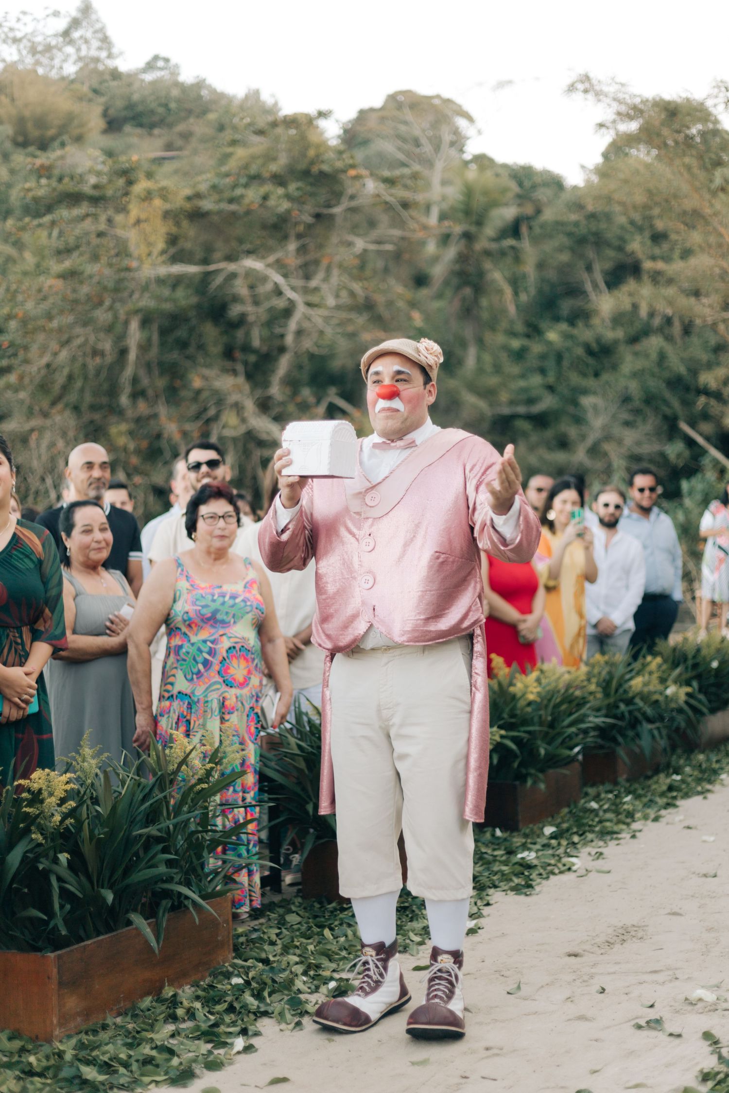 Performer in pink jacket and clown nose entertains wedding guests on an outdoor path surrounded by garden planters.