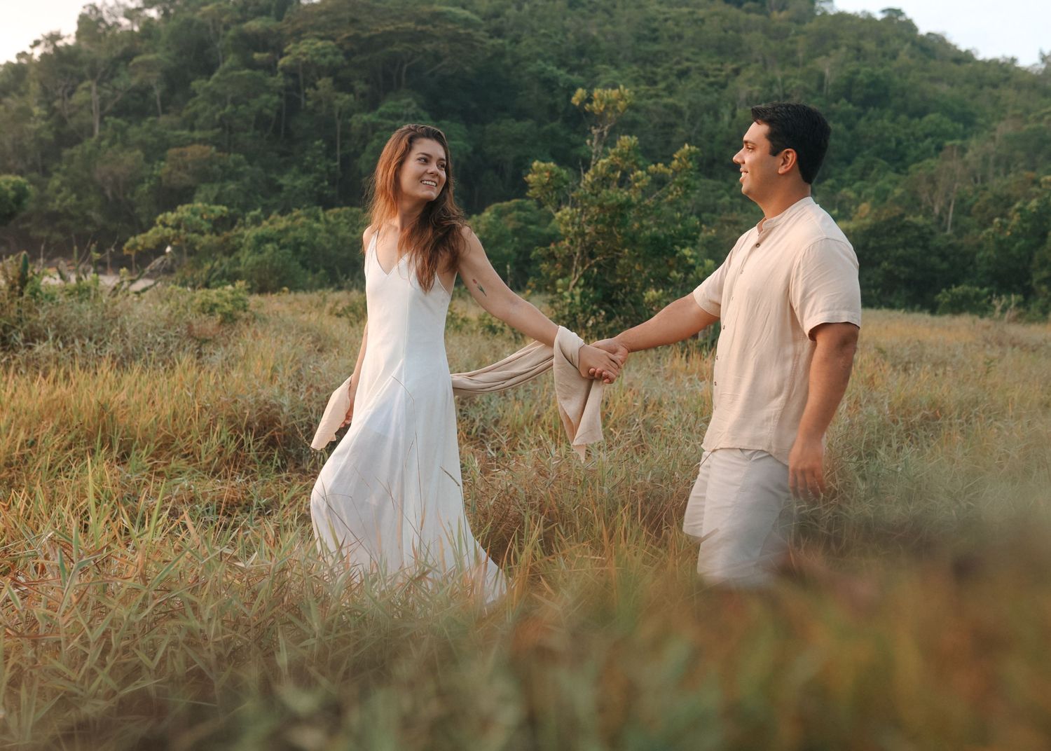 Couple holds hands while walking through tall grass with mountains in the background.