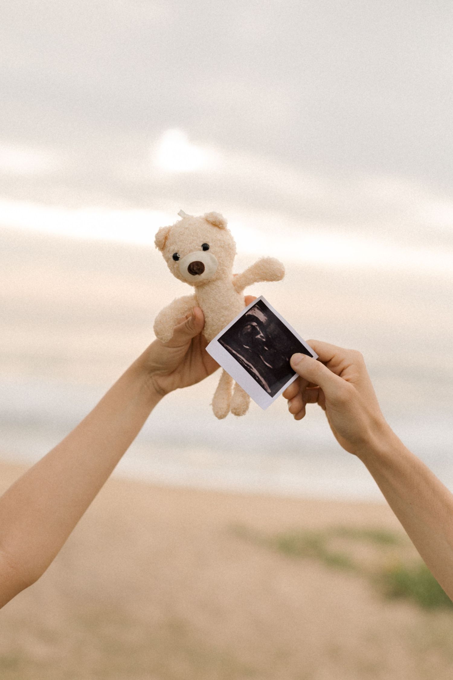 Hands holding up a small teddy bear and ultrasound photo against a sunset sky.