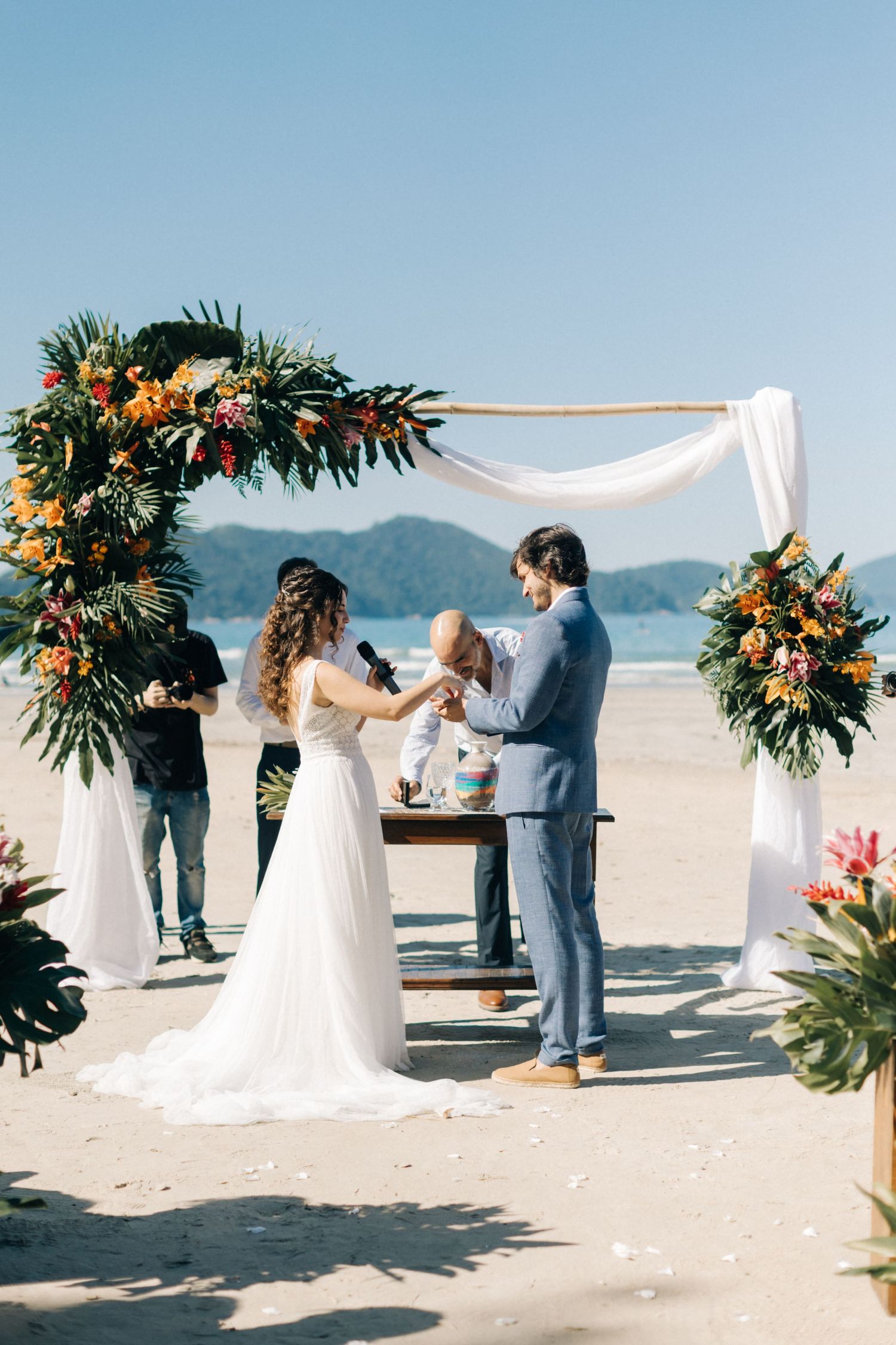 Beach wedding ceremony setup with tropical flower arch and white fabric overlooking mountains.
