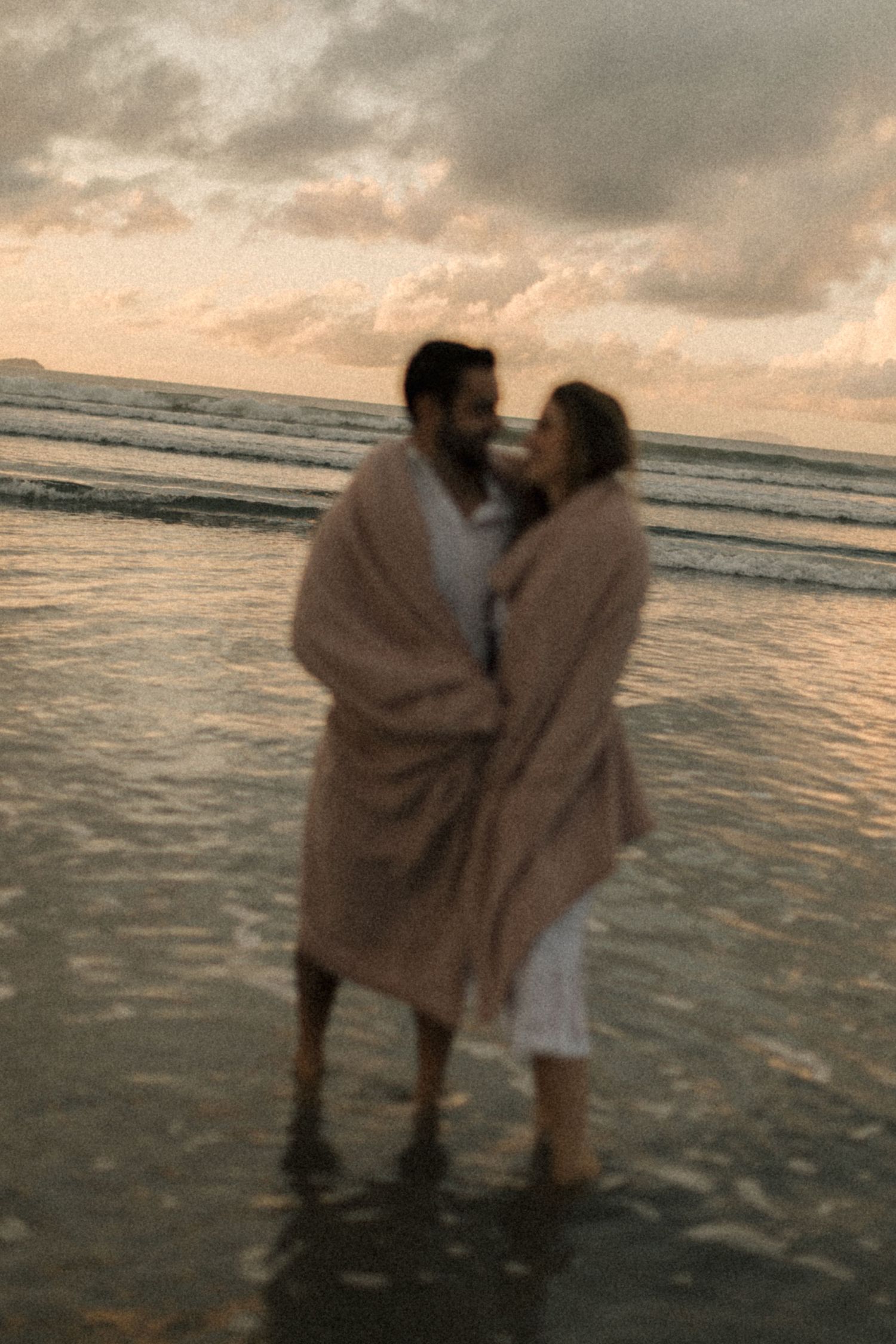 Couple wrapped in blanket embraces on beach at dusk with waves in background.