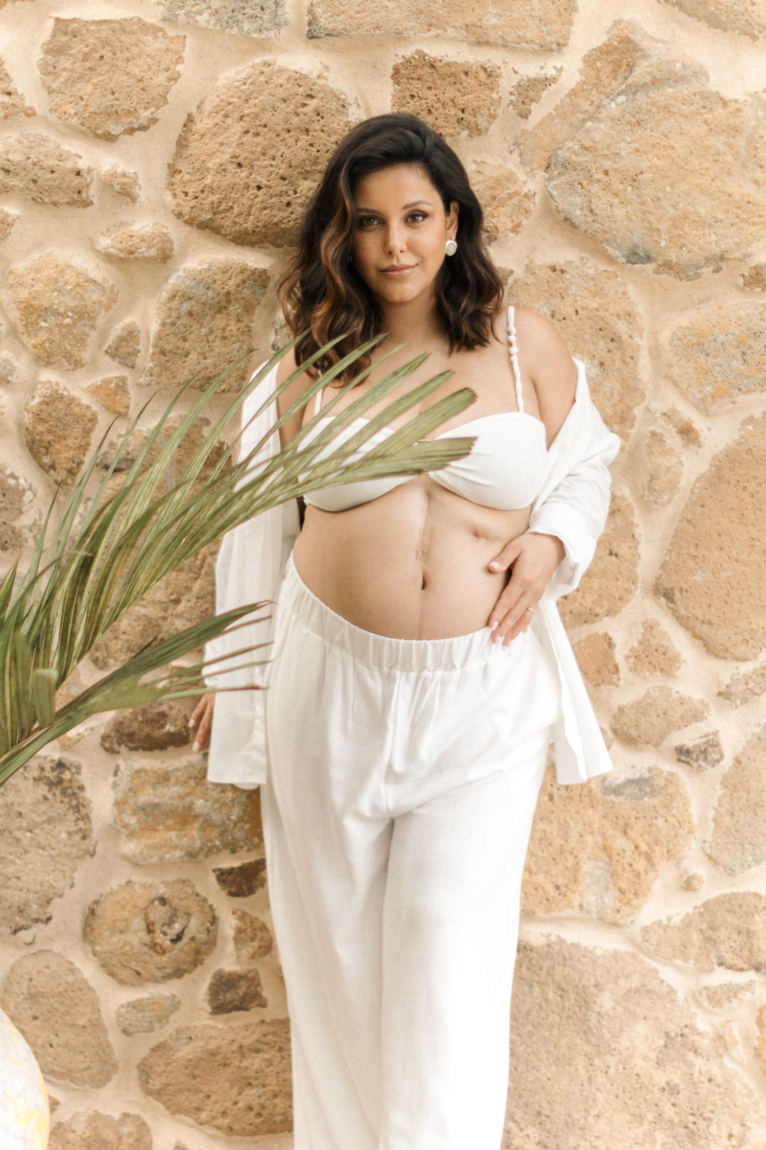 Fashion portrait with palm frond against stone wall featuring white loungewear and crop top in warm natural lighting.