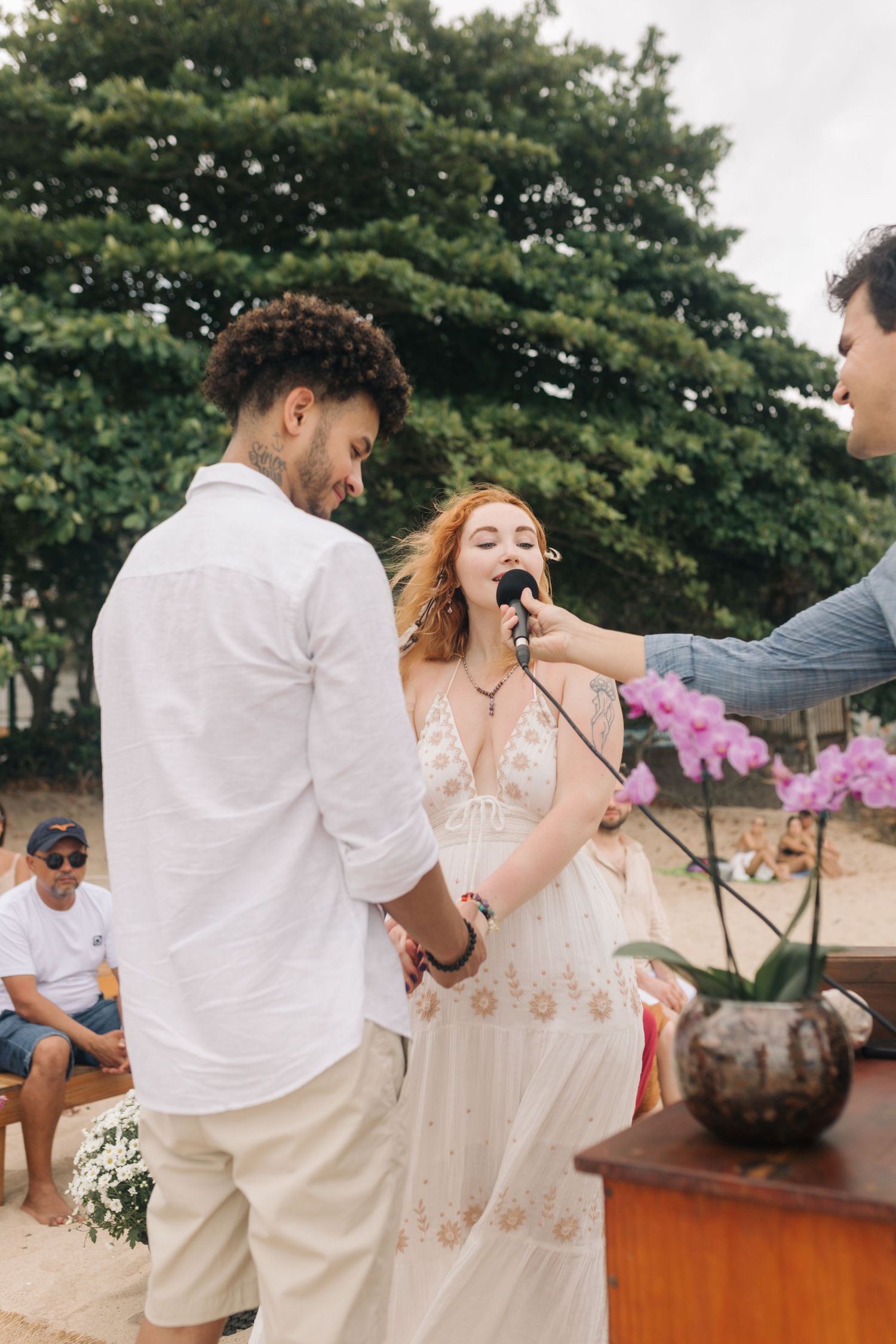 Wedding ceremony on a beach with guests watching as vows are exchanged near blooming orchids.