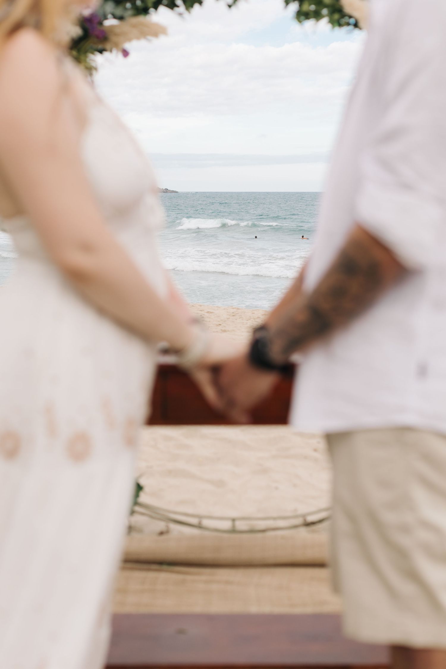 Close-up of couple's hands during beach wedding ceremony with ocean view in background.