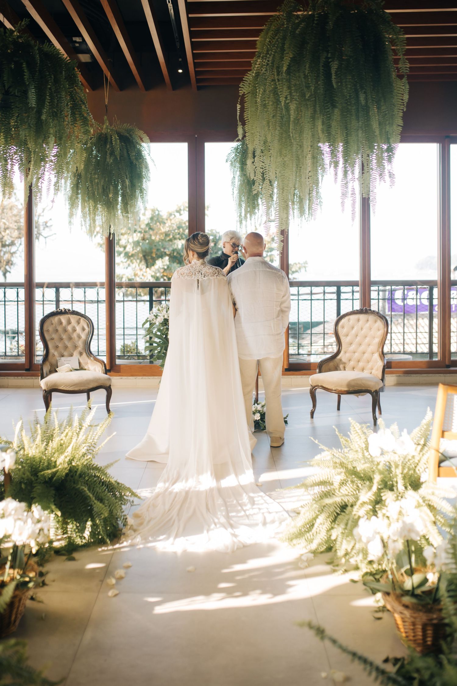 Elegant wedding venue with hanging greenery, vintage chairs, and natural light streaming through windows.