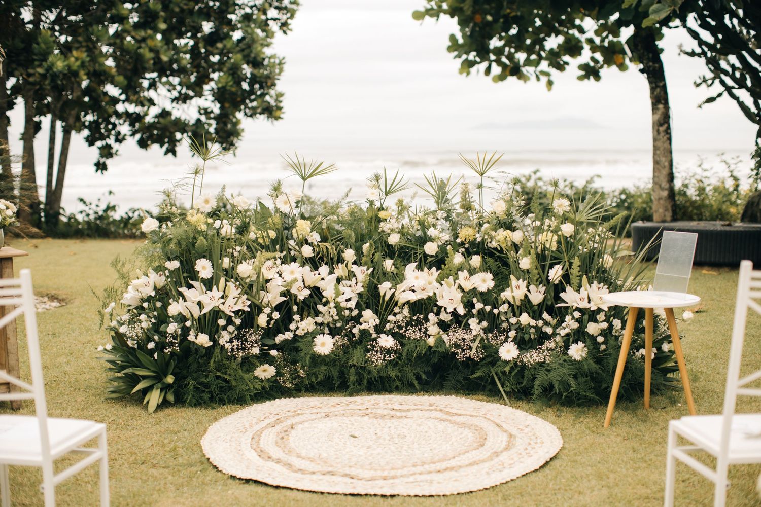 Outdoor wedding ceremony setup with white floral arch and round rug on grass.
