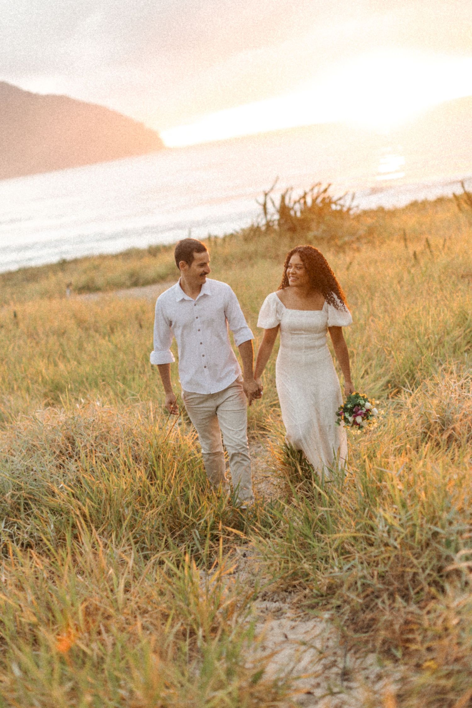 A couple walks hand in hand through golden grass at sunset on a coastal hillside while wearing light summer attire.