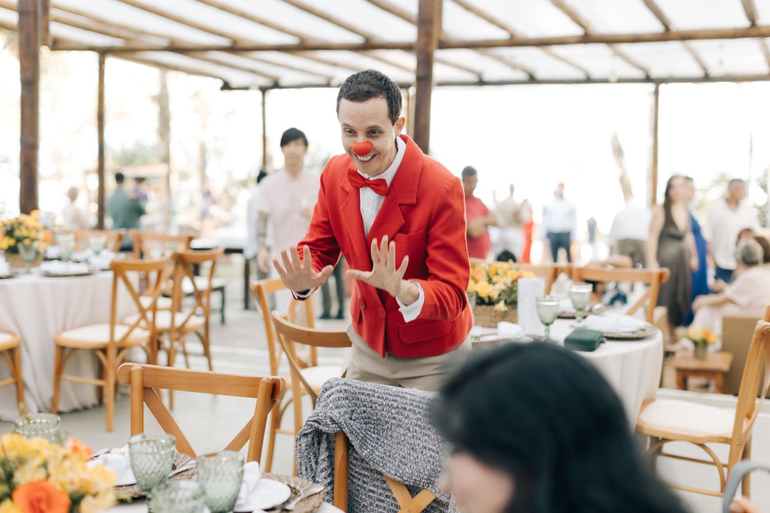 Wedding reception with person in red cardigan gesturing enthusiastically among dining tables.