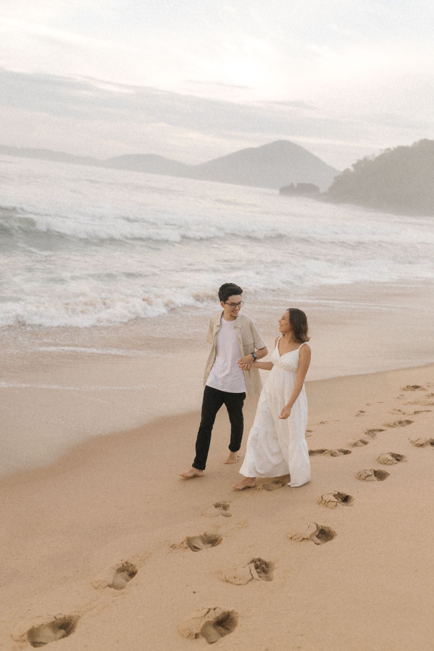 A couple walks hand in hand leaving footprints at the beach during sunset as waves crash on the shore.