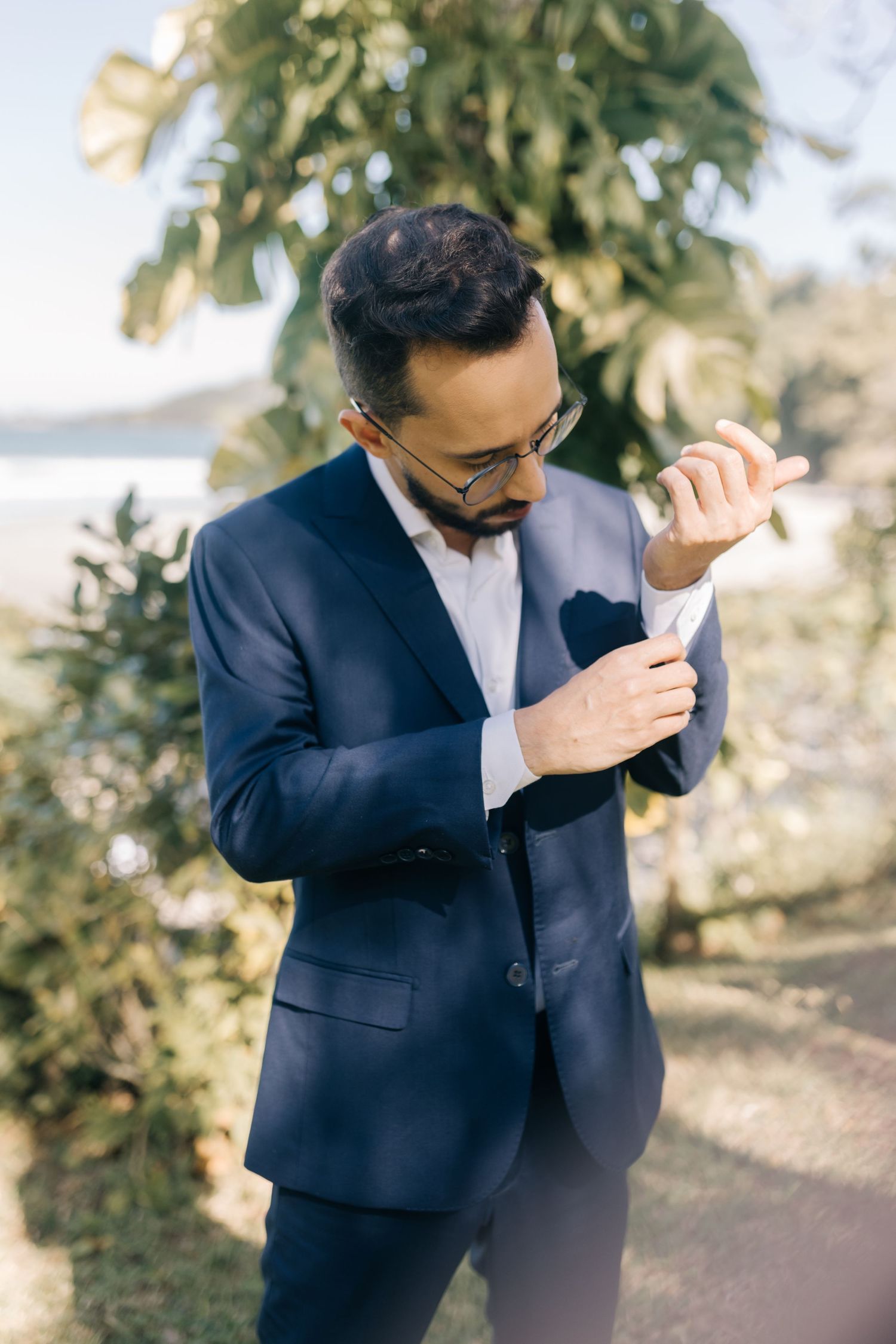 Person in navy suit adjusts sleeve cuff outdoors near coastal vegetation.