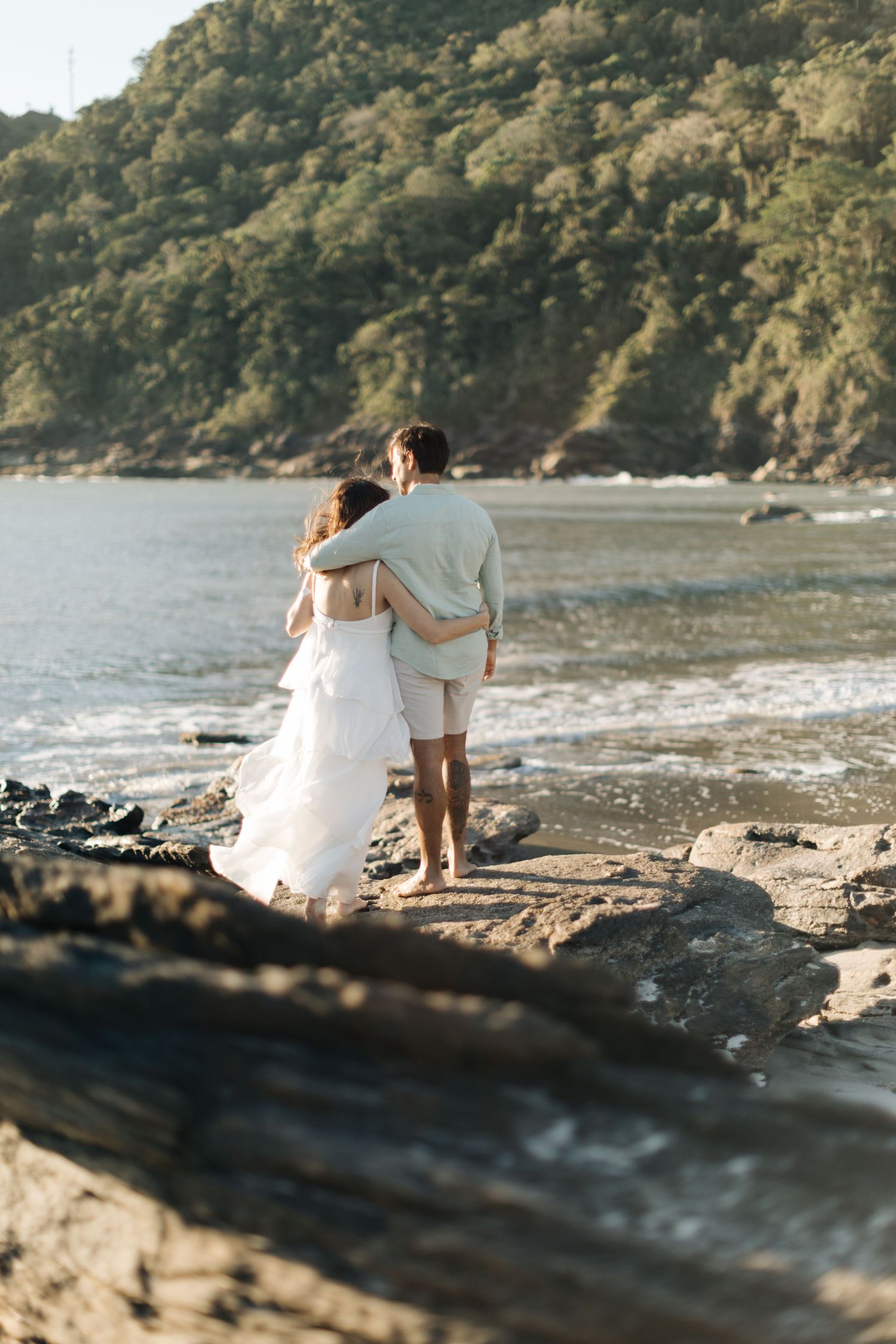 Couple embracing while standing on rocky beach overlooking the ocean.