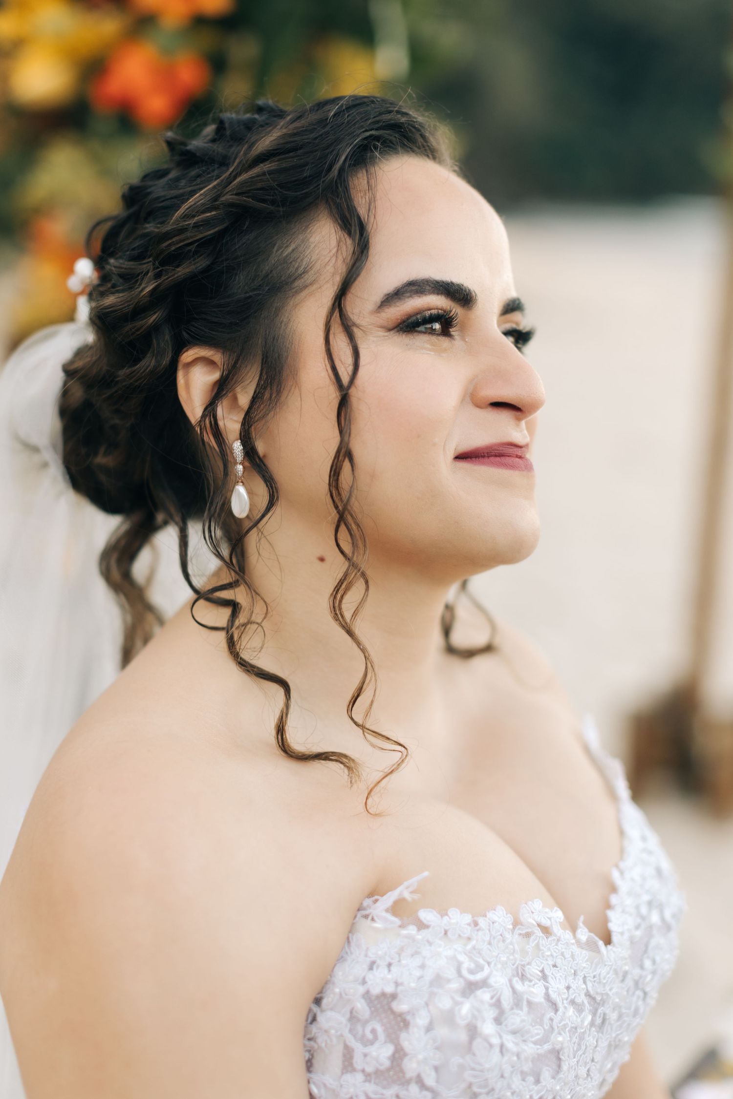 Bride with curled updo and lace dress smiles during beachside wedding.