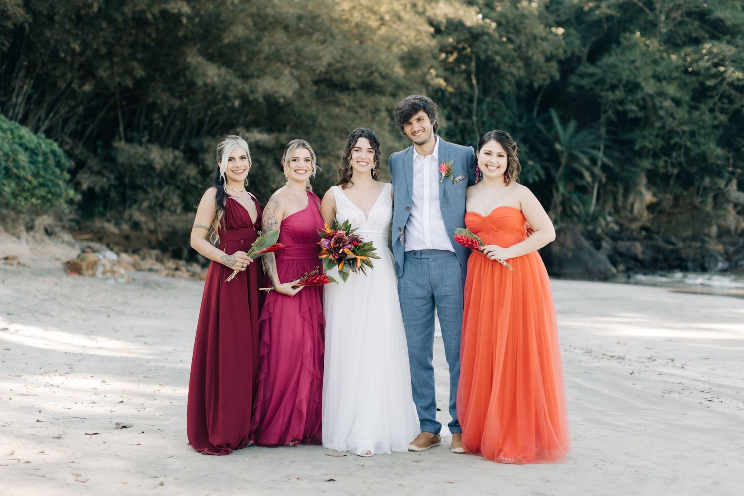 Bridal party in burgundy and orange dresses pose together on a beach.