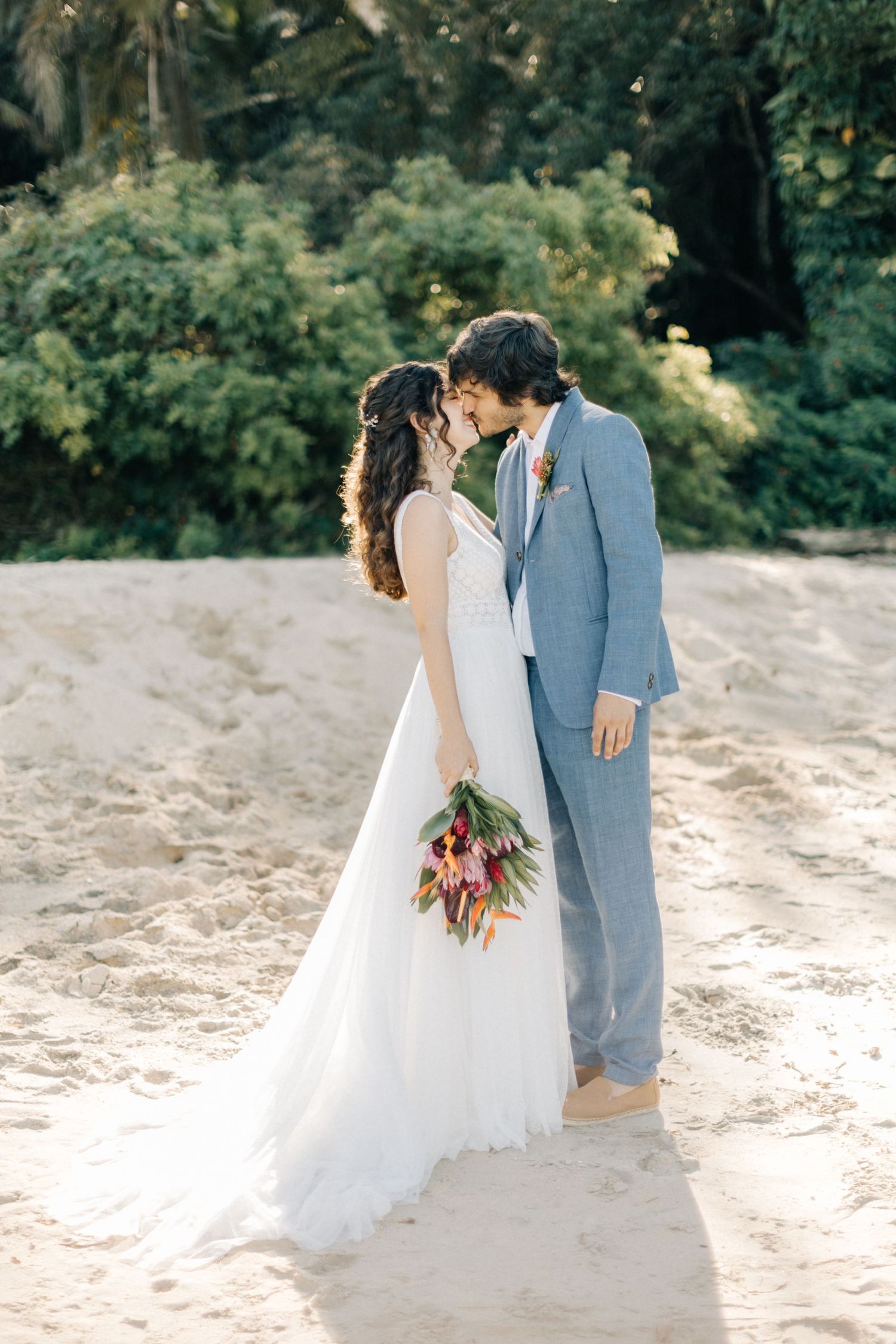 A romantic beach wedding moment with a couple in white dress and light blue suit sharing an intimate kiss.