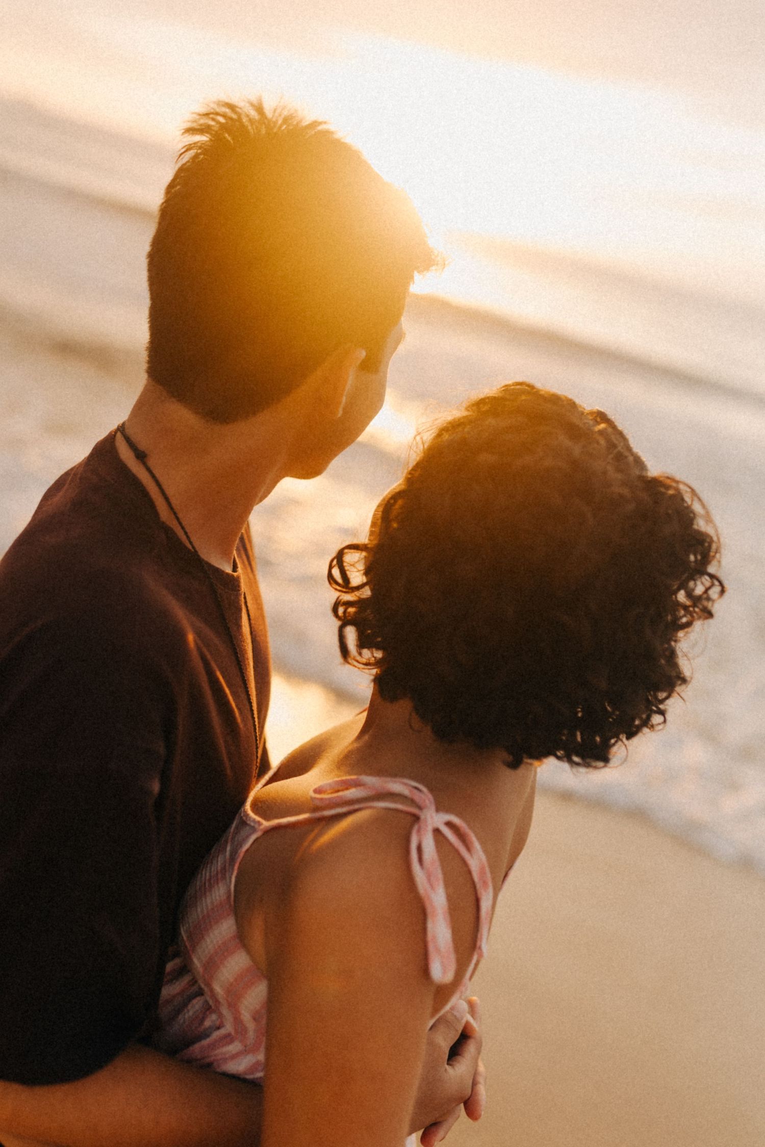 Um casal se abraça durante o pôr do sol da hora dourada na praia, recortado contra o céu quente e brilhante.