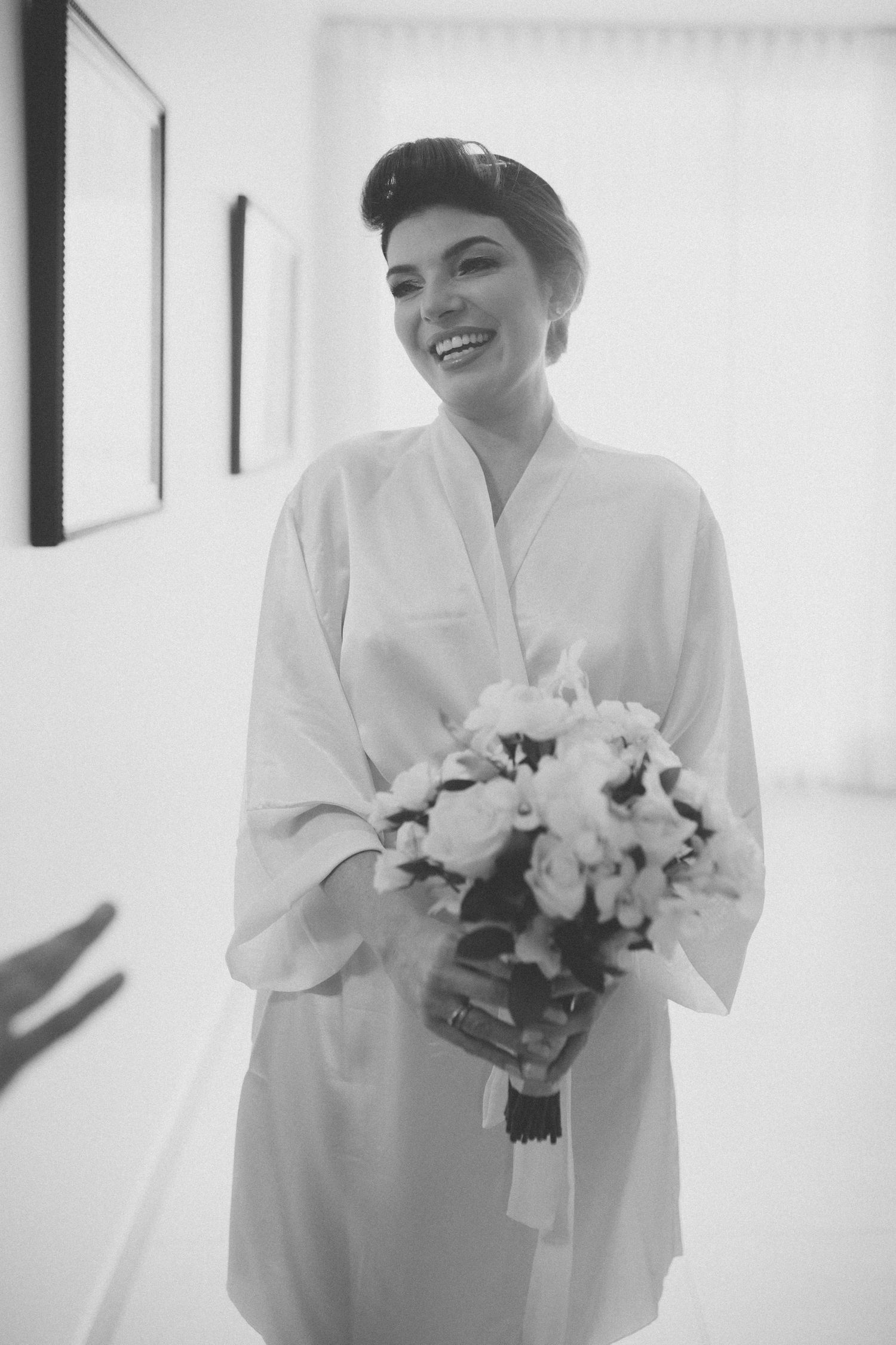 A smiling bride in a white robe holds a bouquet of white flowers during wedding preparations.