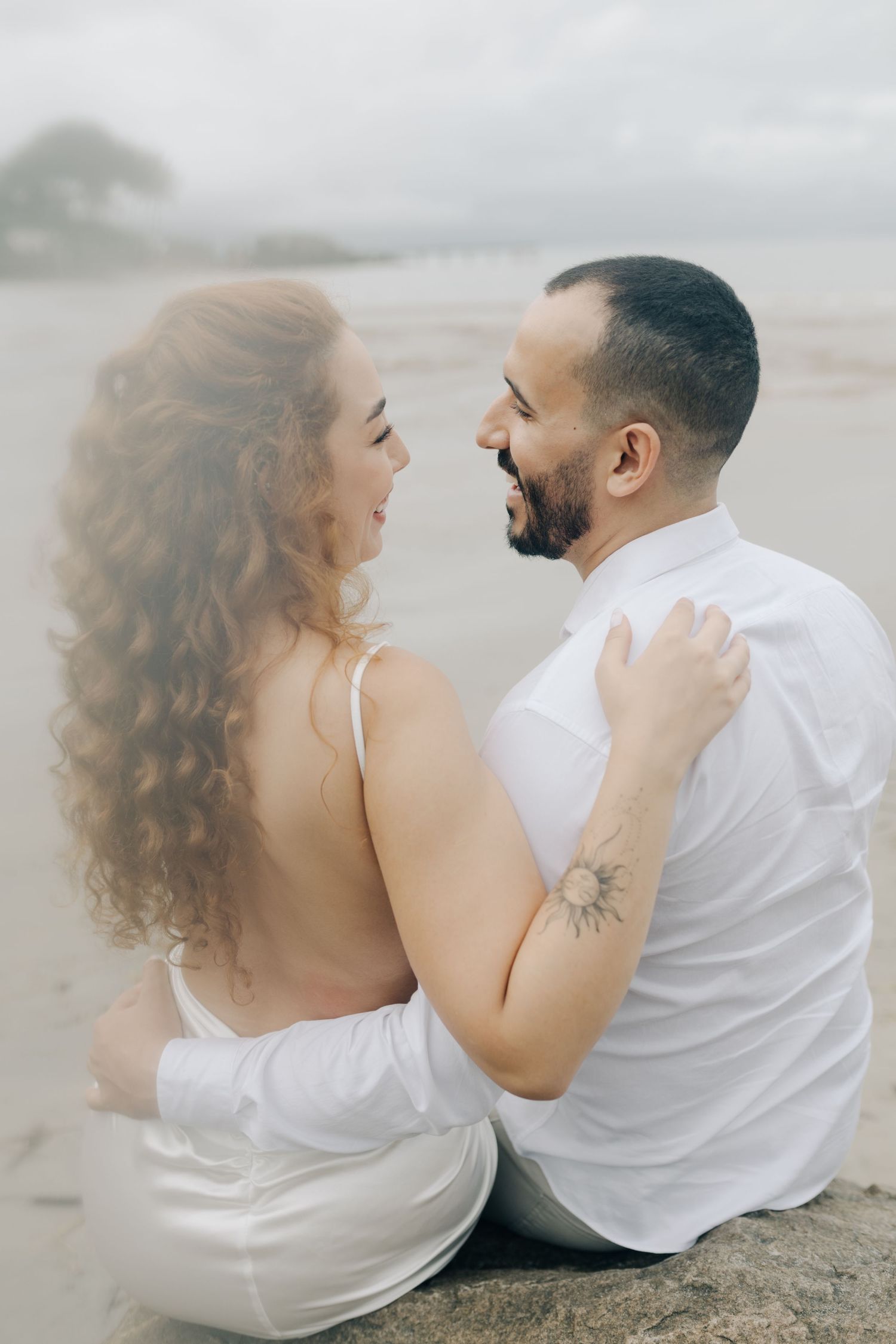 A couple in white clothing shares intimate moments while sitting on a beach during a cloudy day.