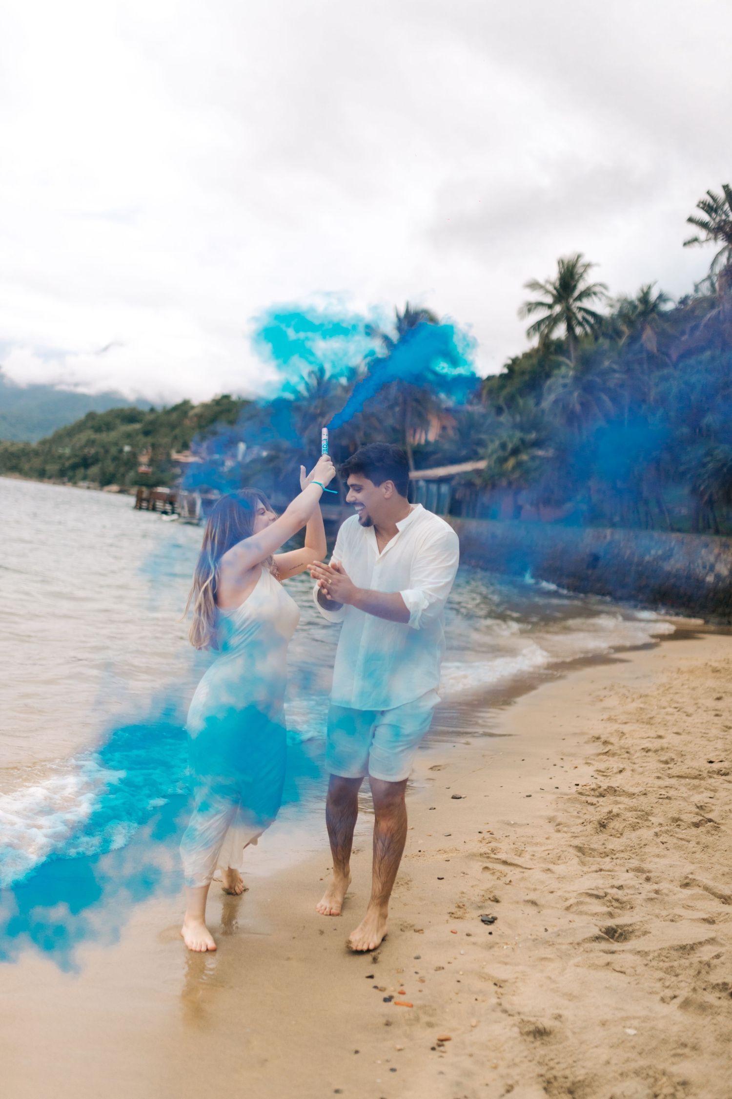 Two people pose with vibrant blue smoke bombs on a tropical beach with palm trees and mountains in the background.