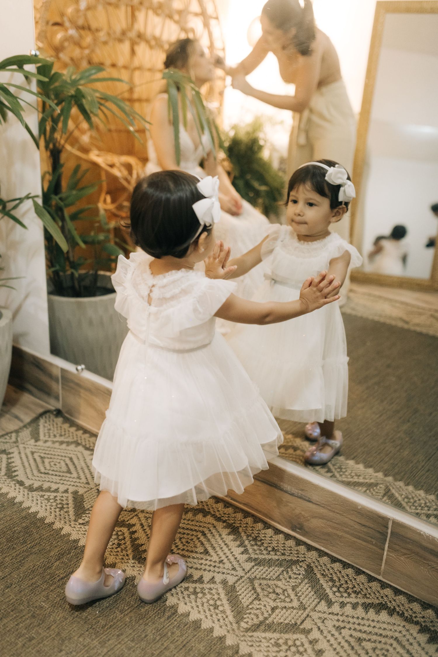 Two flower girls in white dresses and bows playing in front of mirror at wedding venue.