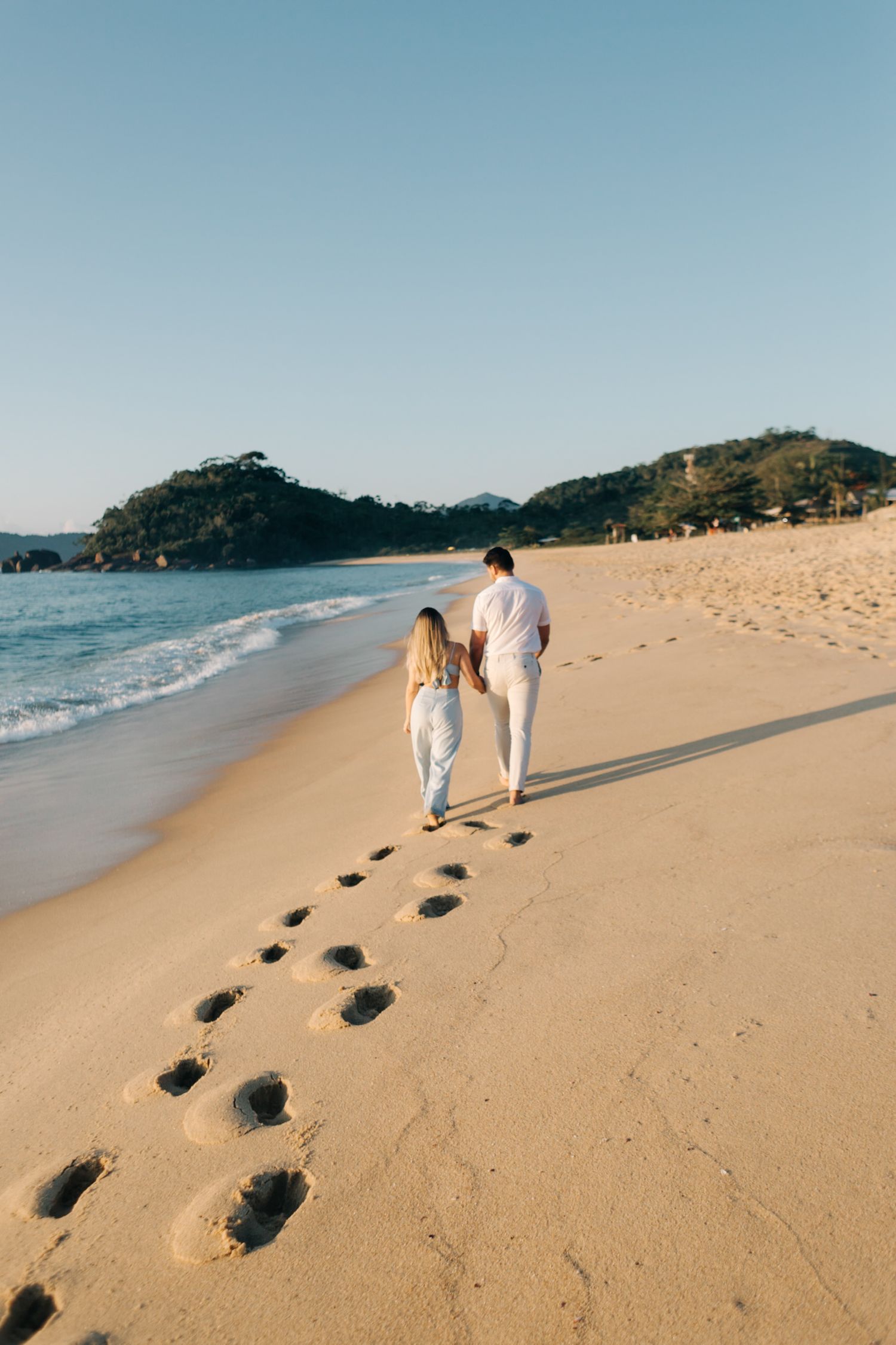 Footprints trail behind a couple walking along a scenic beach with waves and hills in the distance.