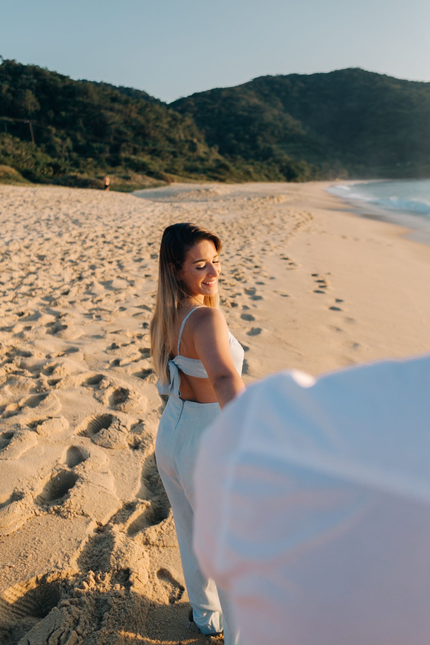 Woman in white two-piece outfit smiling on sandy beach with mountains in background.
