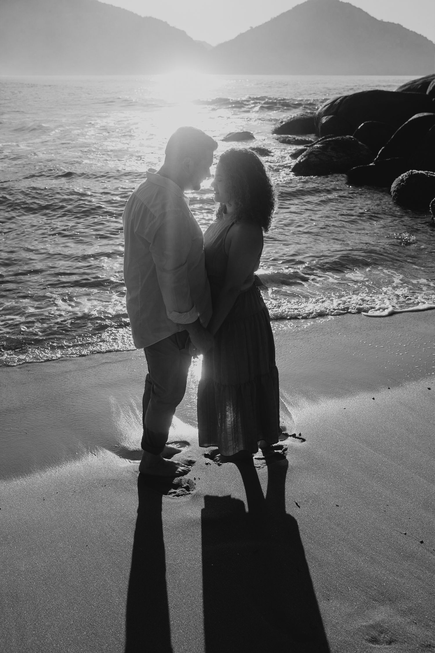 Silhouette of couple standing close together on rocky beach with mountains and ocean waves.