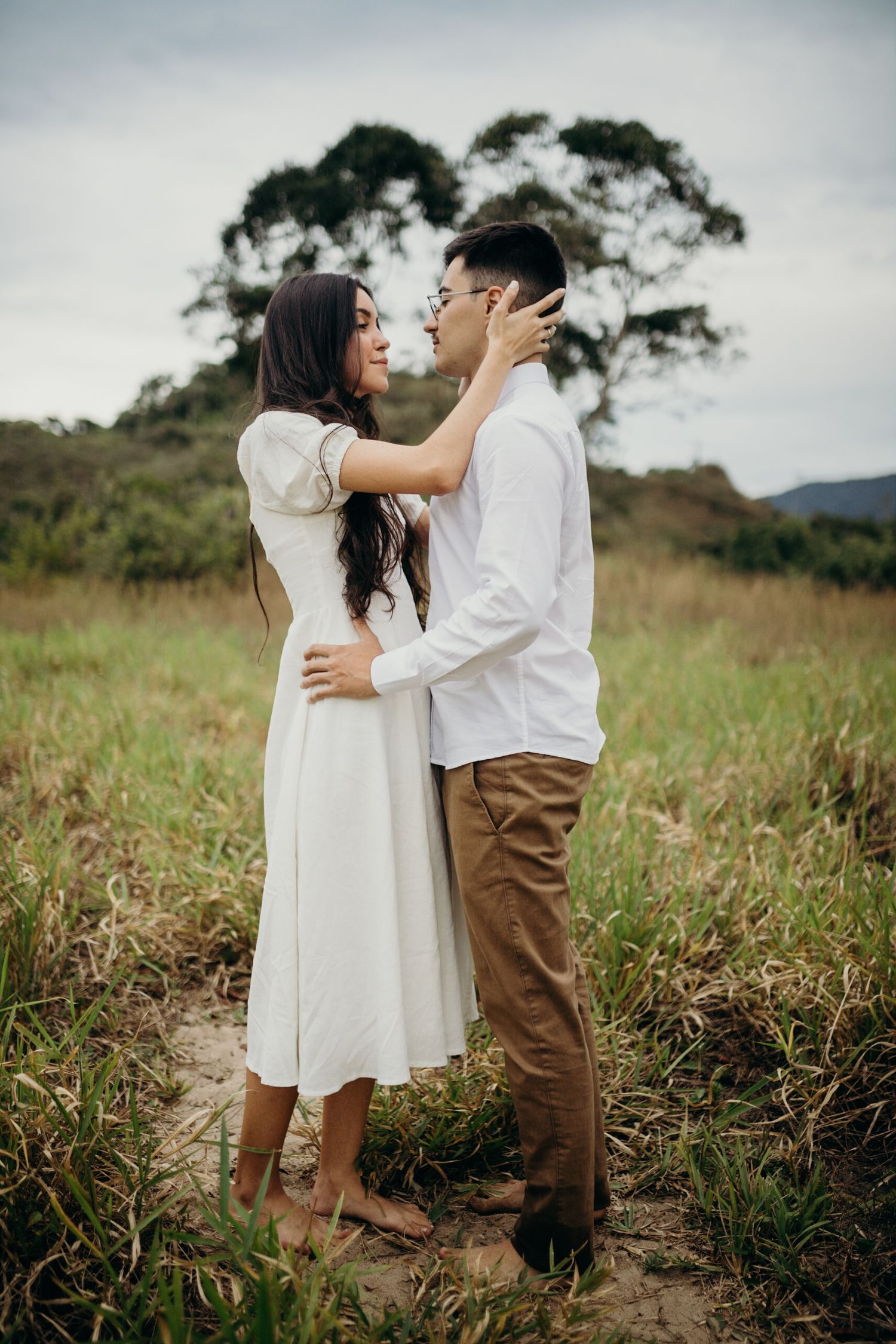 A couple embracing tenderly in a field with eucalyptus trees and mountains behind them.