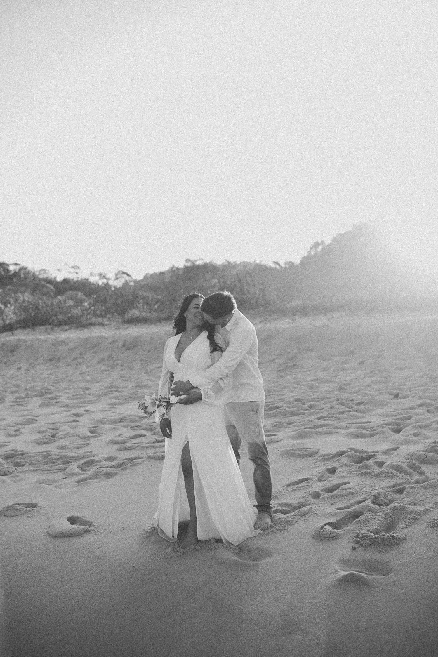 Black and white beach photography captures intimate moments between couple in flowing white clothing.