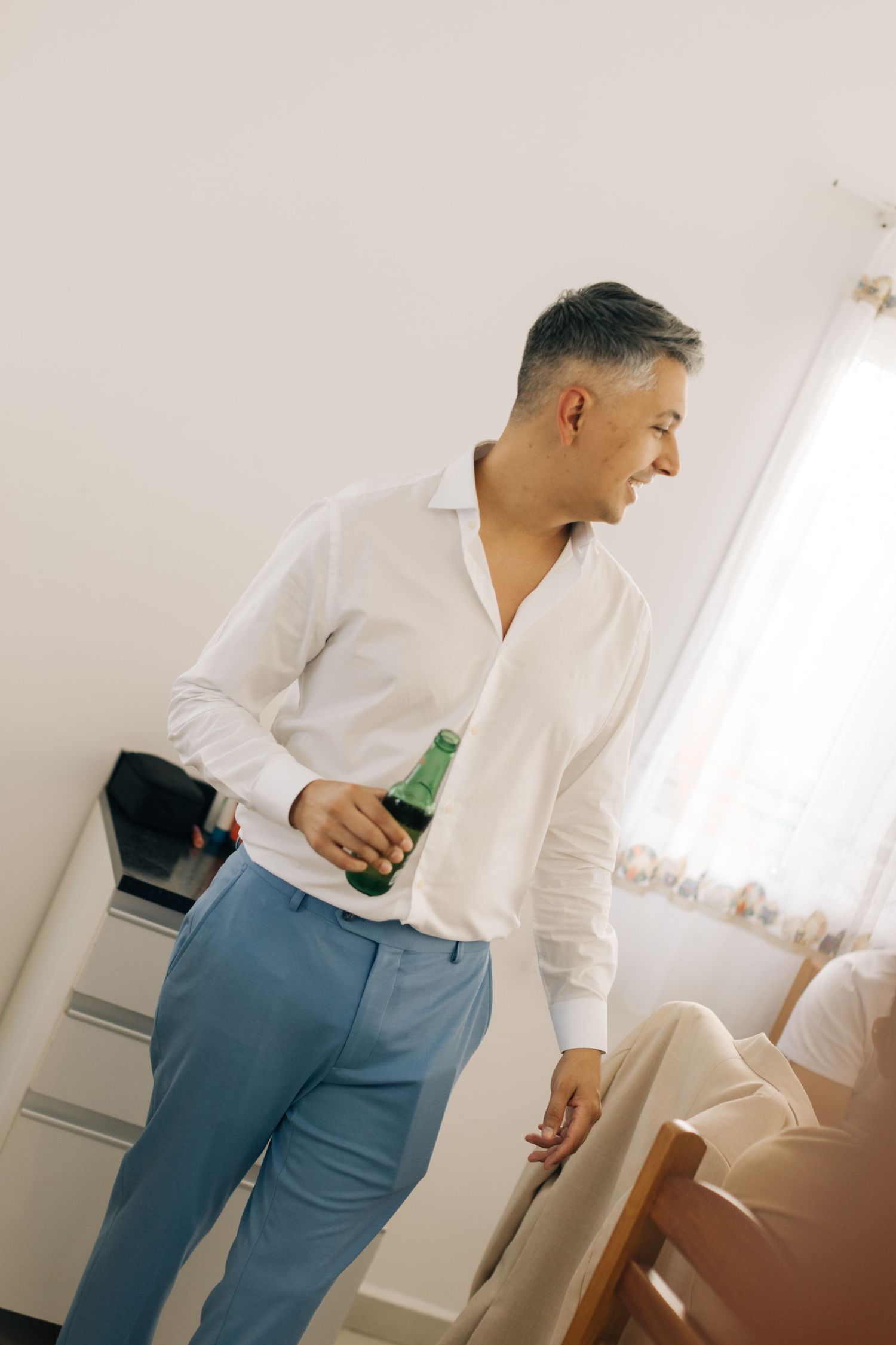 Man in crisp white shirt and blue trousers holding a beverage in bright room.