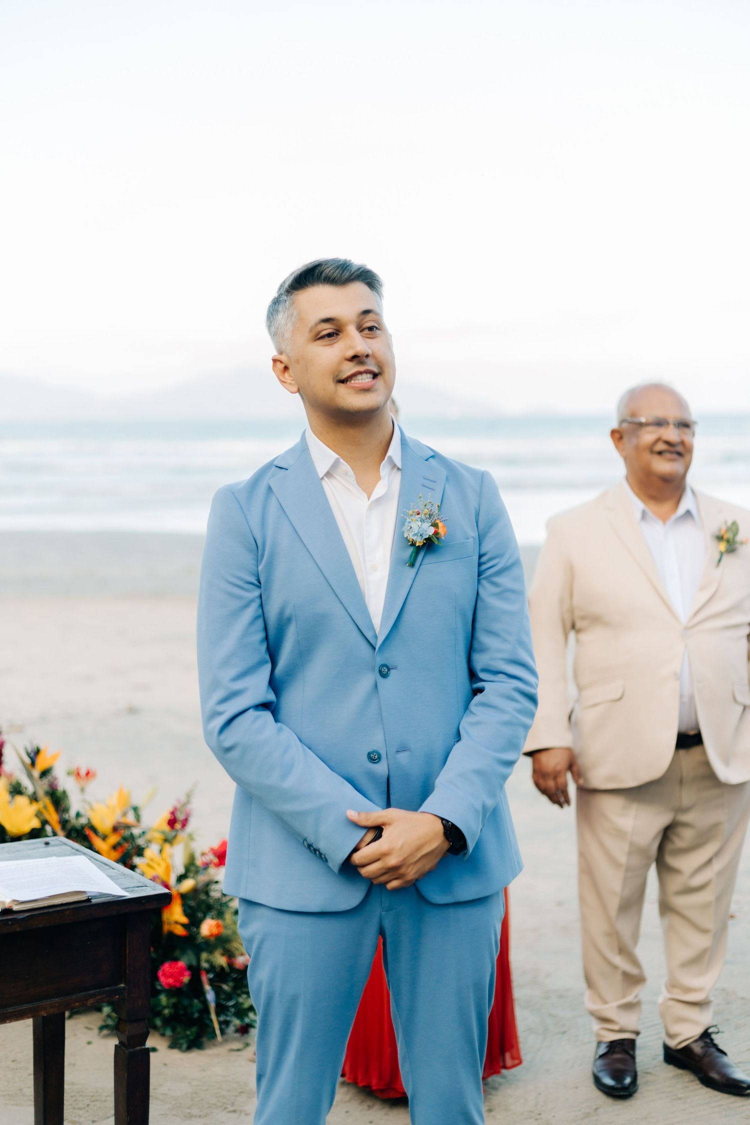 A light blue suited man smiles at a beach wedding ceremony with yellow flowers and ocean backdrop.