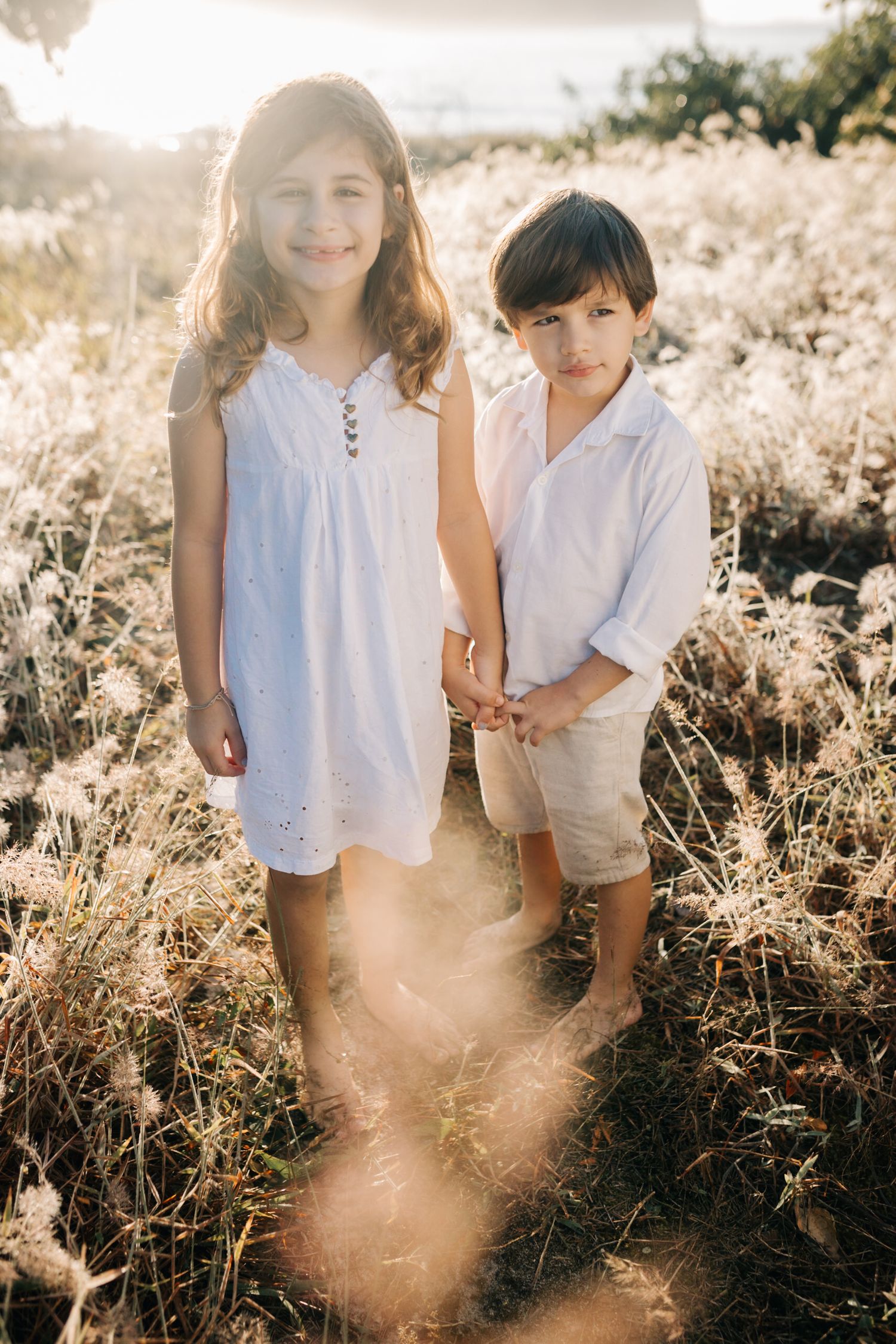 Two children stand together in a sunlit field wearing white and beige summer clothing.
