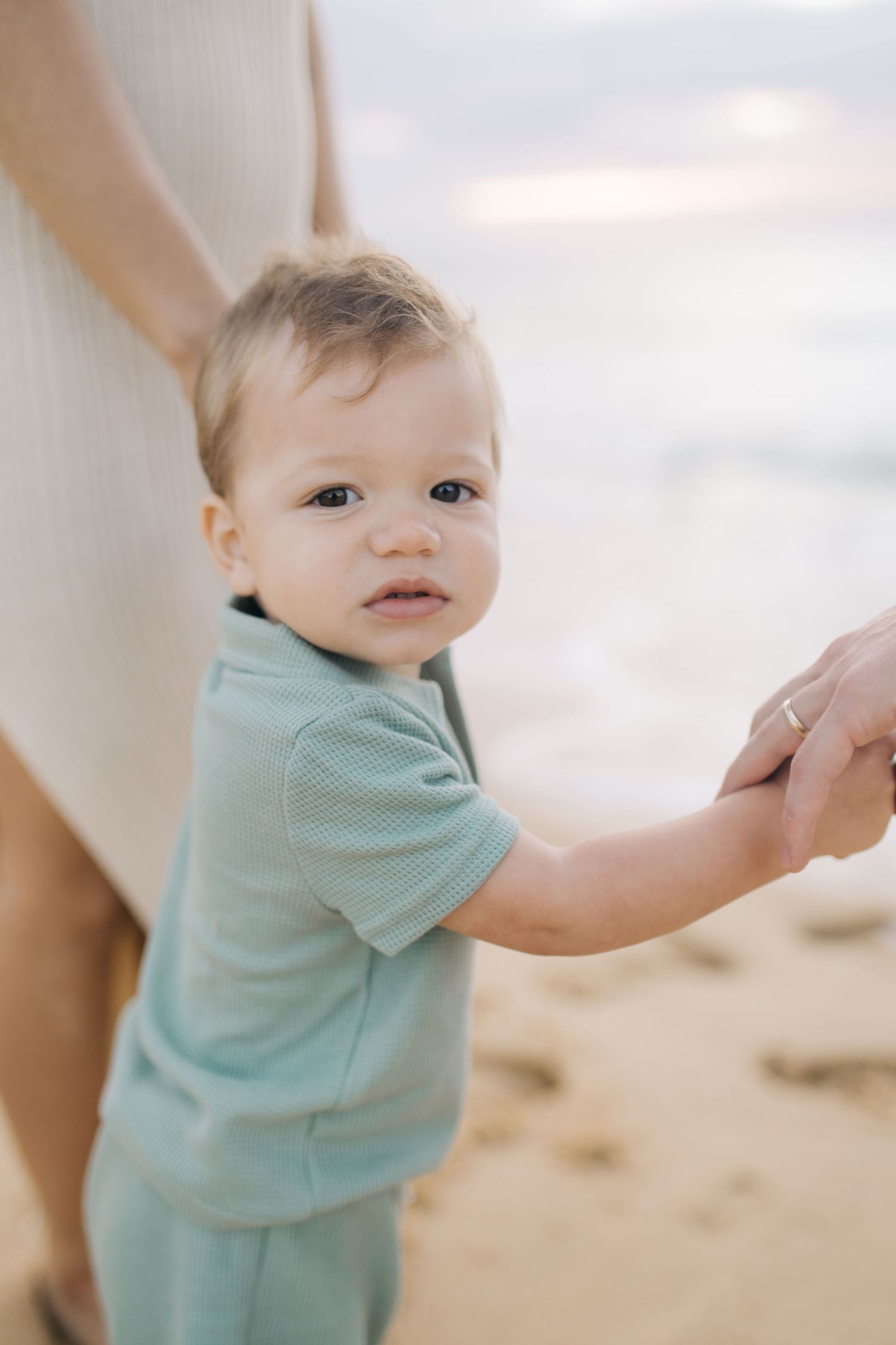 A toddler in light blue clothing holds hands with someone on a beach.